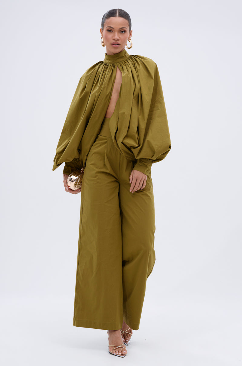 A woman wears the GOLD COAST BALLOON SLEEVE BLOUSE—an olive green, long-sleeve top with dramatic gathered fabric and a front keyhole cutout—styled with matching wide-leg trousers, gold hoop earrings, a gold clutch, and strappy heels.