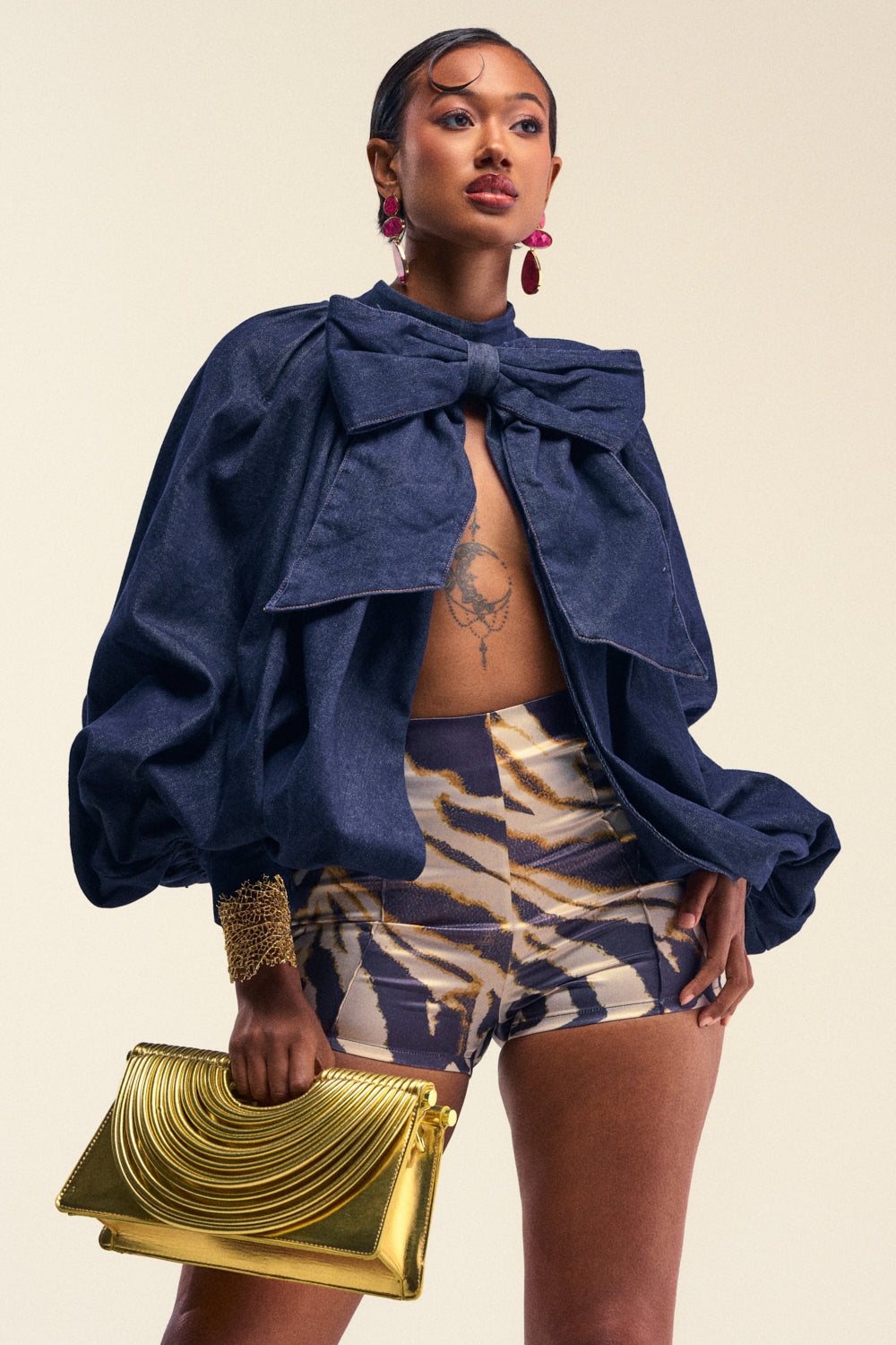 A woman poses confidently in the LEAH RUCHED DENIM MOCK NECK TOP IN DARK BLUE DENIM, styled under a striking blue oversized jacket with a large bow. She pairs it with patterned shorts, accessorized by a gold clutch, gold bracelet, and red earrings. Her slicked-back hair and bold makeup complete the look.