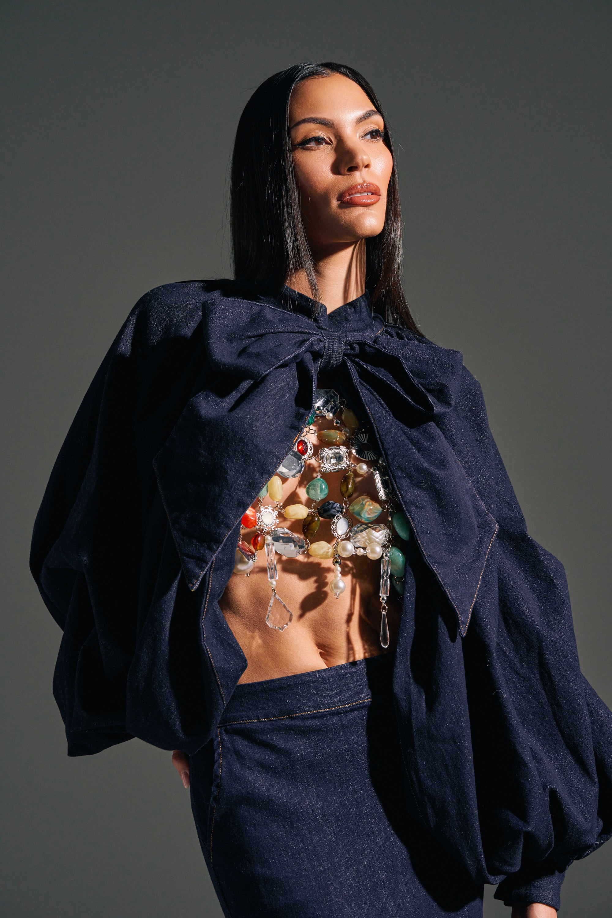 A woman with long dark hair confidently poses against a plain background, wearing a voluminous dark jacket with a bold bow over the LEAH RUCHED DENIM MOCK NECK TOP IN DARK BLUE DENIM and a matching dark skirt.