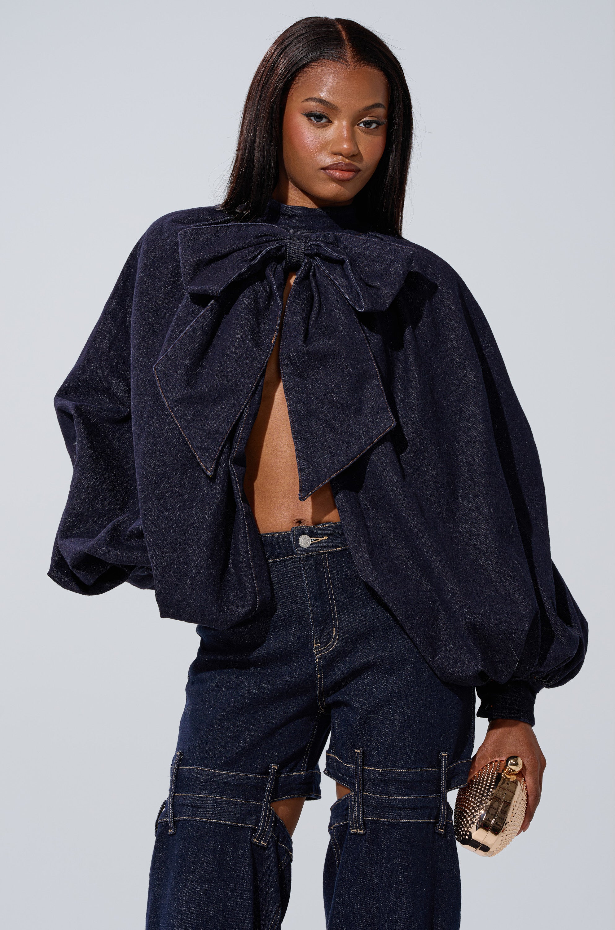 A woman poses confidently in the LEAH Ruched Denim Mock Neck Top in dark blue denim, featuring dramatic balloon sleeves and a large bow, partially open at the front. She pairs it with high-waisted pants and a textured clutch.