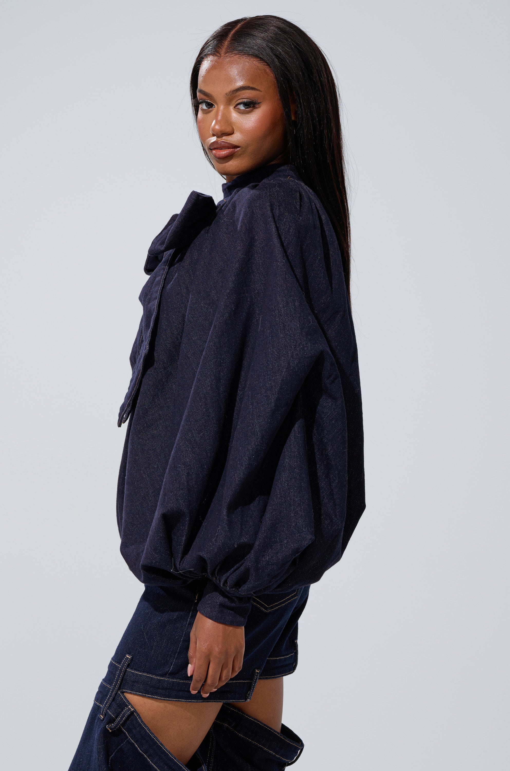 A woman with long straight hair wears the LEAH Ruched Denim Mock Neck Top in dark blue denim, paired with jeans featuring cut-out sections, standing against a plain light background.