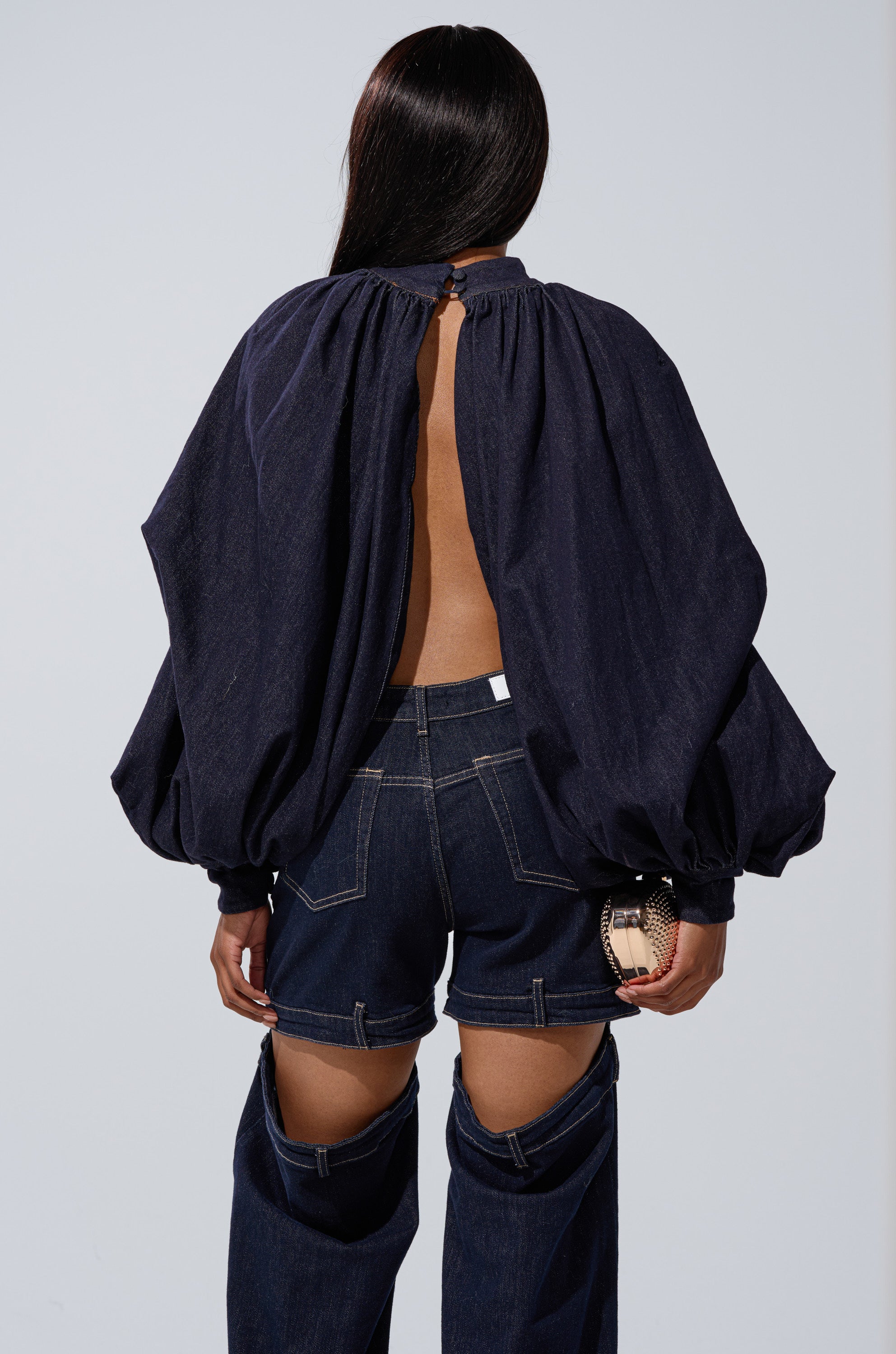 A woman, seen from behind, wears the LEAH RUCHED DENIM MOCK NECK TOP IN DARK BLUE DENIM with voluminous sleeves and an open back, paired with dark blue cutout jeans. She carries a small round clutch.