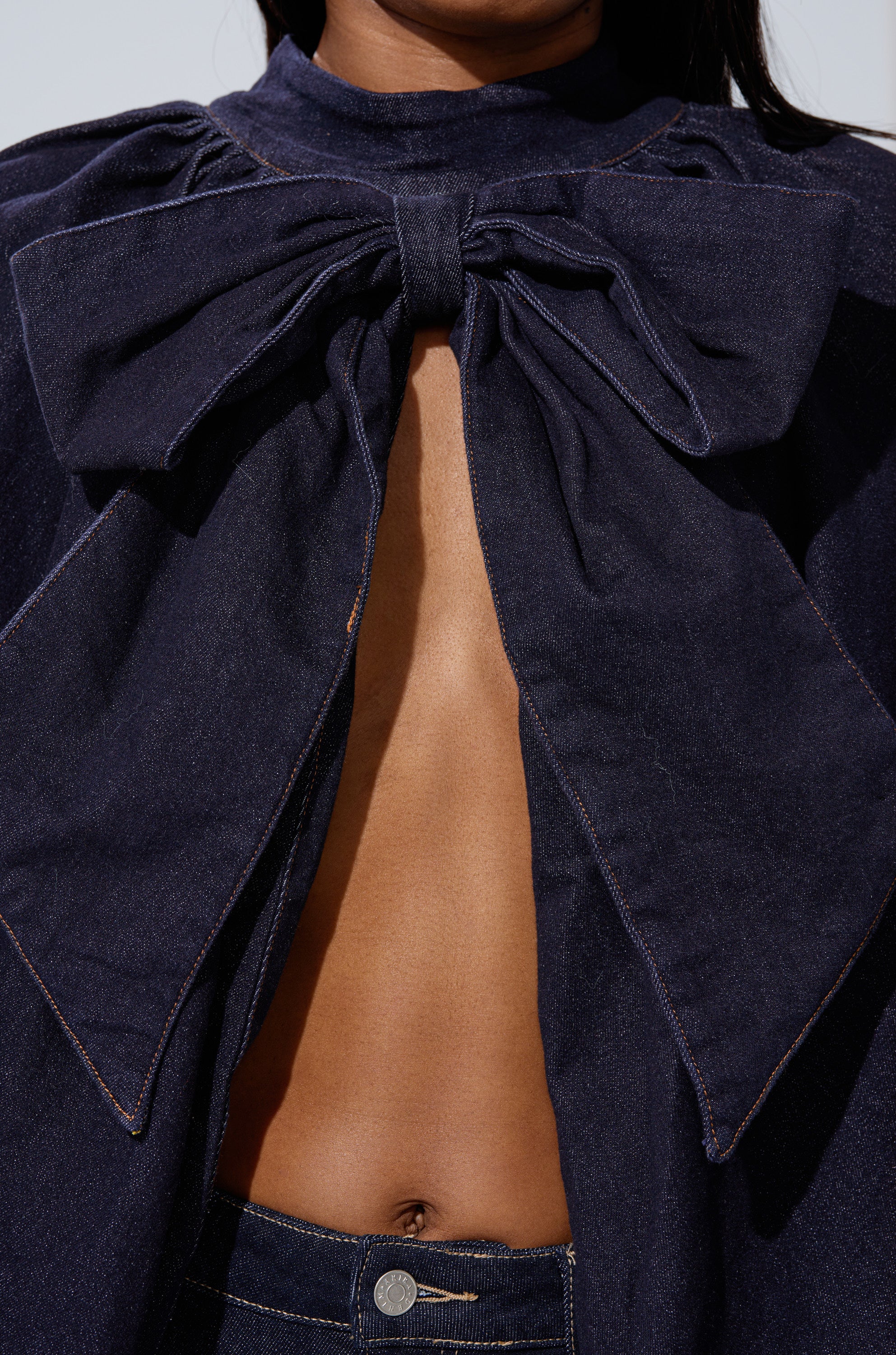 A person wearing the LEAH Ruched Denim Mock Neck Top in dark blue denim, featuring a large bow at the chest, open front showing the midriff, paired with high-waisted jeans.