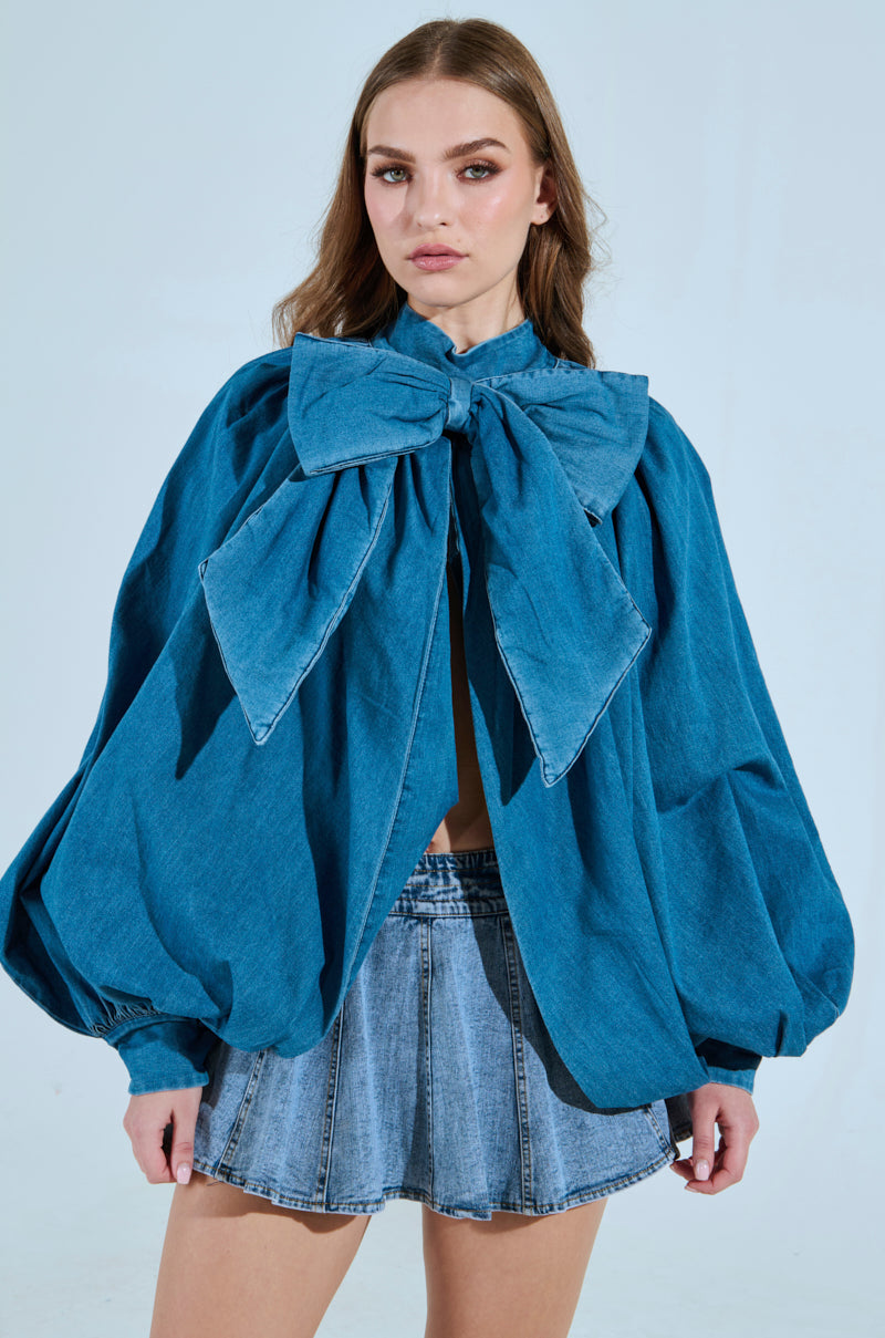 Wearing a MASTERPIECE MEDIUM DENIM LONG SLEEVE PUFF BLOUSE and pleated denim mini skirt, their long brown hair cascades gracefully as they stand against a light background with a neutral expression. 0