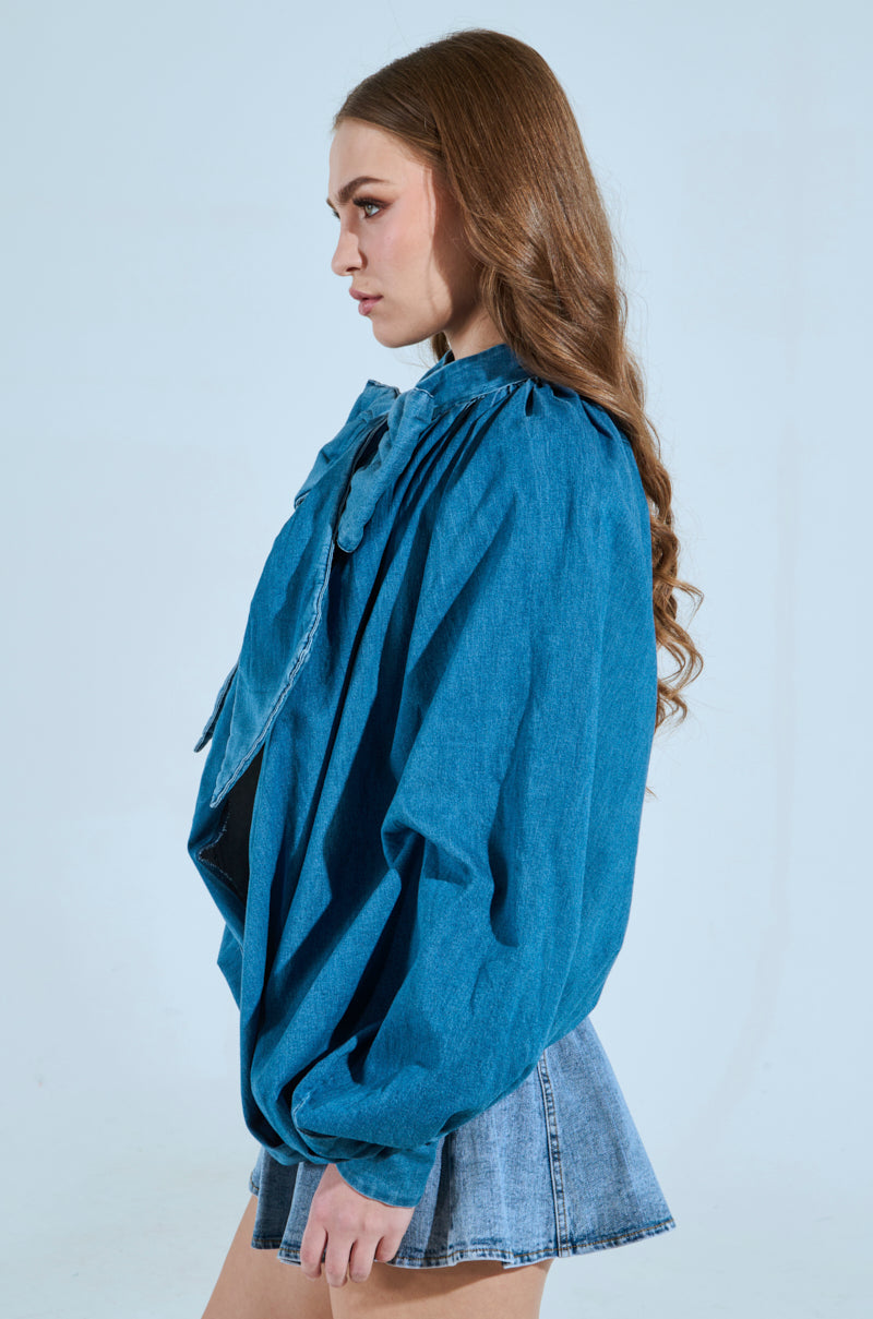 A person with long hair is wearing the Masterpiece Medium Denim Long Sleeve Puff Blouse paired with a faded denim skirt. They are posed against a plain light background, looking to their right. 2