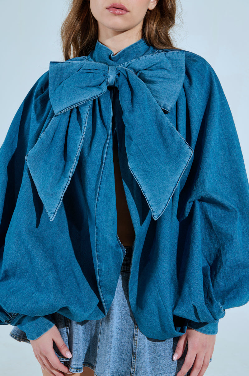 A woman wears the MASTERPIECE Medium Denim Long Sleeve Puff Blouse, featuring an oversized design with a large bow collar and puff sleeves, paired perfectly with a light denim skirt. Her face is partially out of view. 5