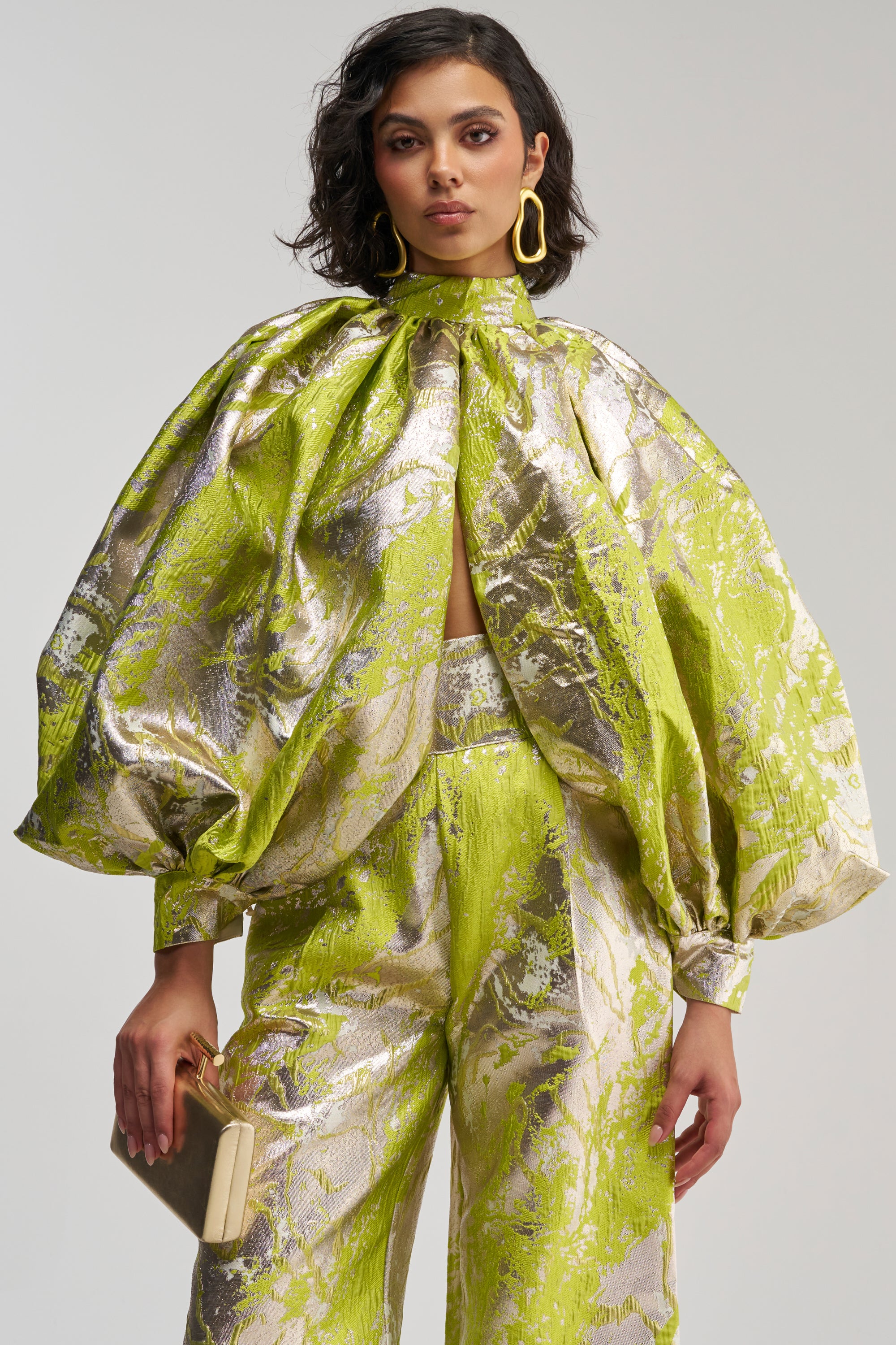 A person wearing the ELECTRIC GARDEN BROCADE TOP, a shiny neon green and silver patterned piece with voluminous sleeves, paired with matching pants, accessorized with yellow earrings and holding a beige clutch.