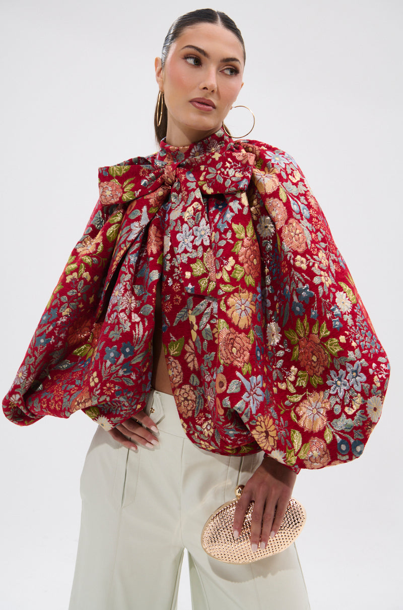 A woman wears the ETHEREAL FLORAL PUFF SLEEVE BROCADE TOP with metallic details, white high-waisted pants, large hoop earrings, and holds a small textured gold clutch against a plain white background.