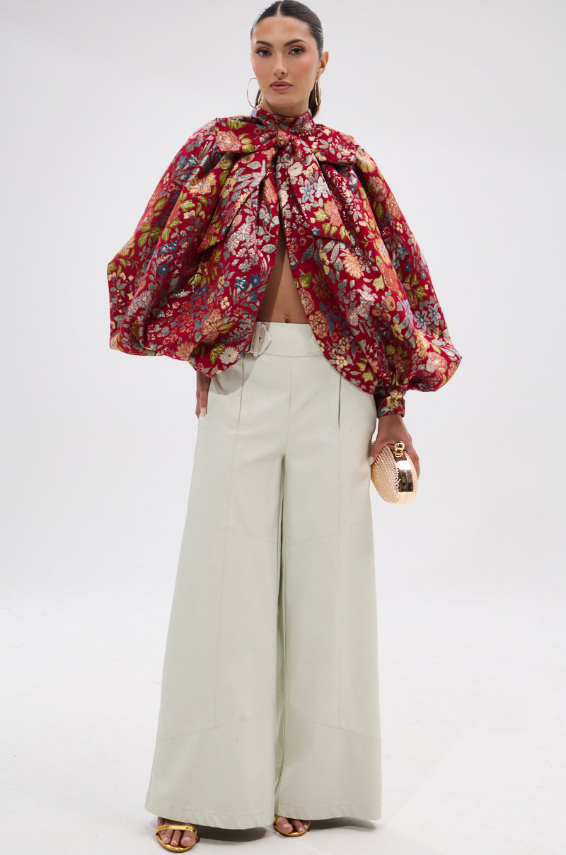 A woman poses in the ETHEREAL FLORAL PUFF SLEEVE BROCADE TOP with a bow neckline and puff sleeves, styled with high-waisted cream wide-leg trousers, gold sandals, hoop earrings, and a textured gold clutch.