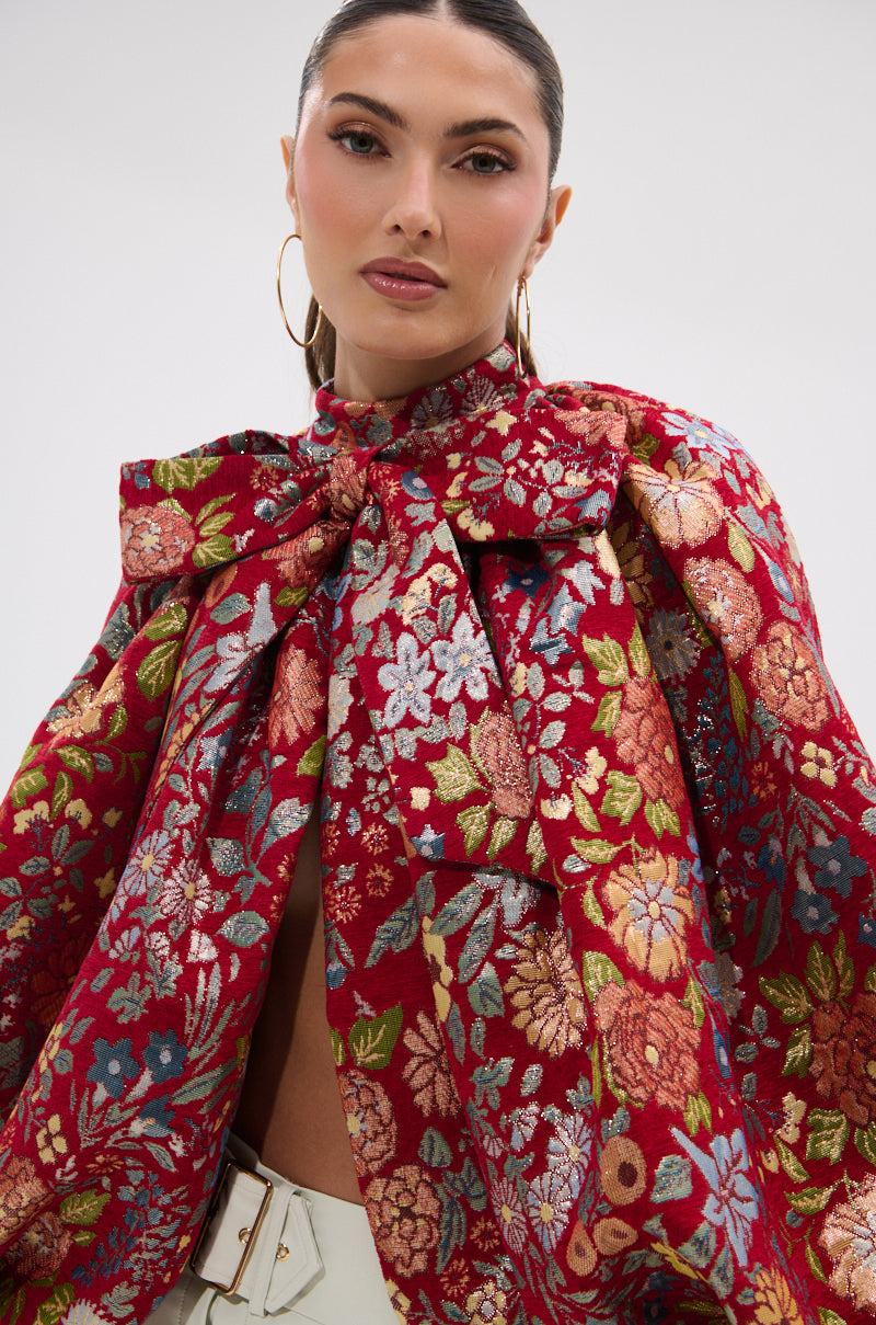 A woman with slicked-back hair and gold hoop earrings wears the ETHEREAL FLORAL PUFF SLEEVE BROCADE TOP, styled as a red cape with a large bow at the neck, paired with light high-waisted pants featuring a wide belt.