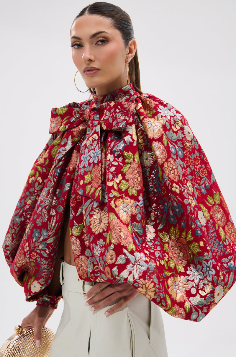 A woman with slicked-back hair wears the ETHEREAL FLORAL PUFF SLEEVE BROCADE TOP, styled as a red cape with a large bow and voluminous sleeves. She holds a gold textured clutch and wears hoop earrings against a plain white background.