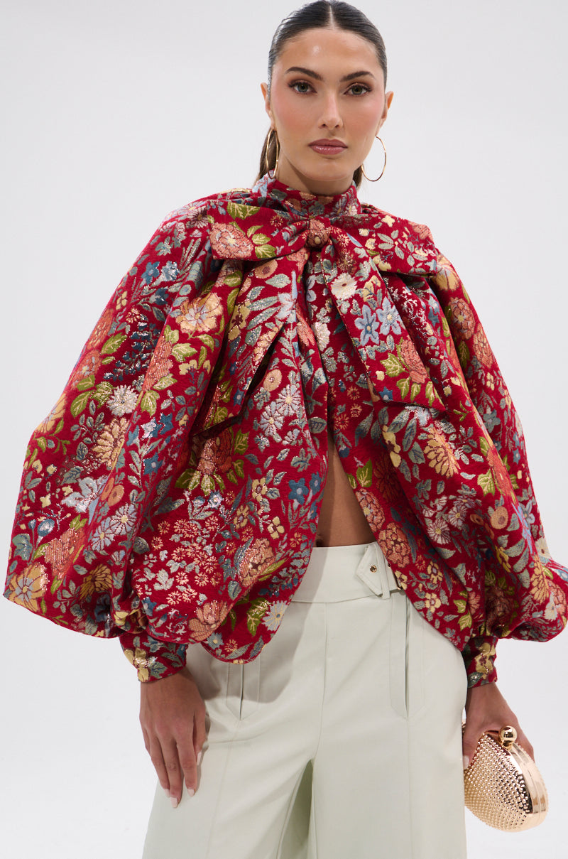 A woman wears the ETHEREAL FLORAL PUFF SLEEVE BROCADE TOP with voluminous sleeves, paired with light high-waisted pants, large hoop earrings, and a small textured gold clutch featuring metallic details.