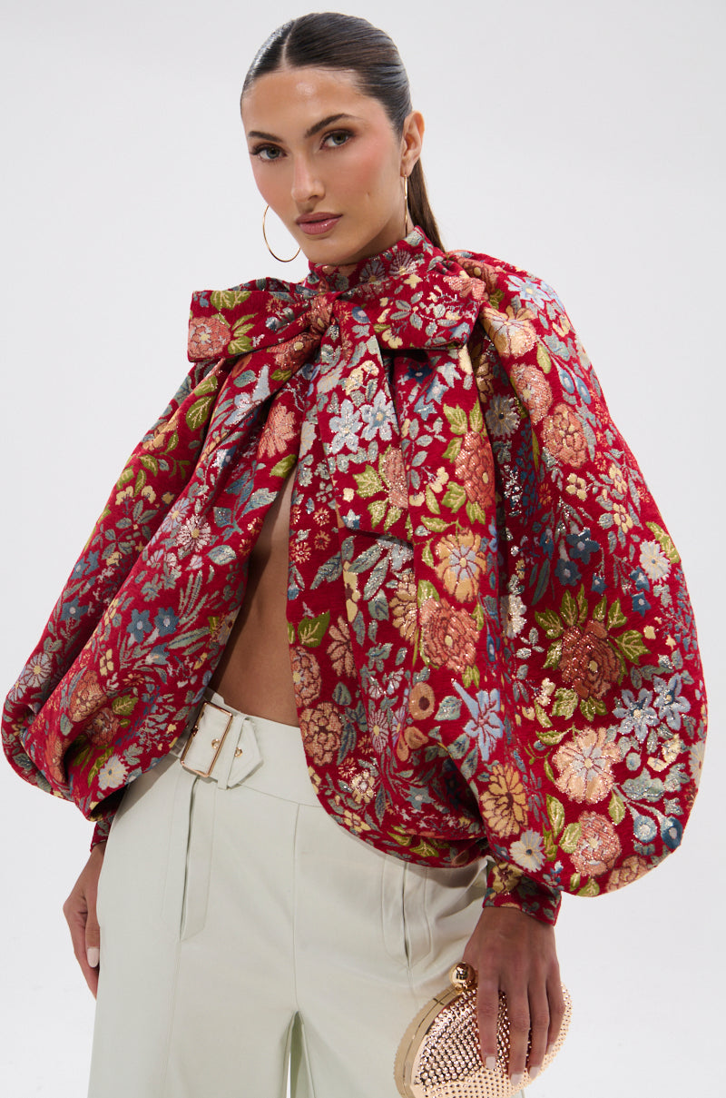 A woman with long dark hair wears the ETHEREAL FLORAL PUFF SLEEVE BROCADE TOP with a large neck bow, white high-waisted pants, hoop earrings, and holds a gold clutch against a plain background.