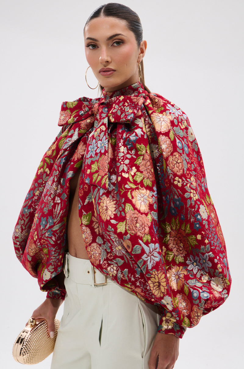 A woman with slicked-back hair wears large hoop earrings, high-waisted white pants, and the ETHEREAL FLORAL PUFF SLEEVE BROCADE TOP with a big bow at the neck. She finishes her look with a gold textured clutch featuring metallic detailing.