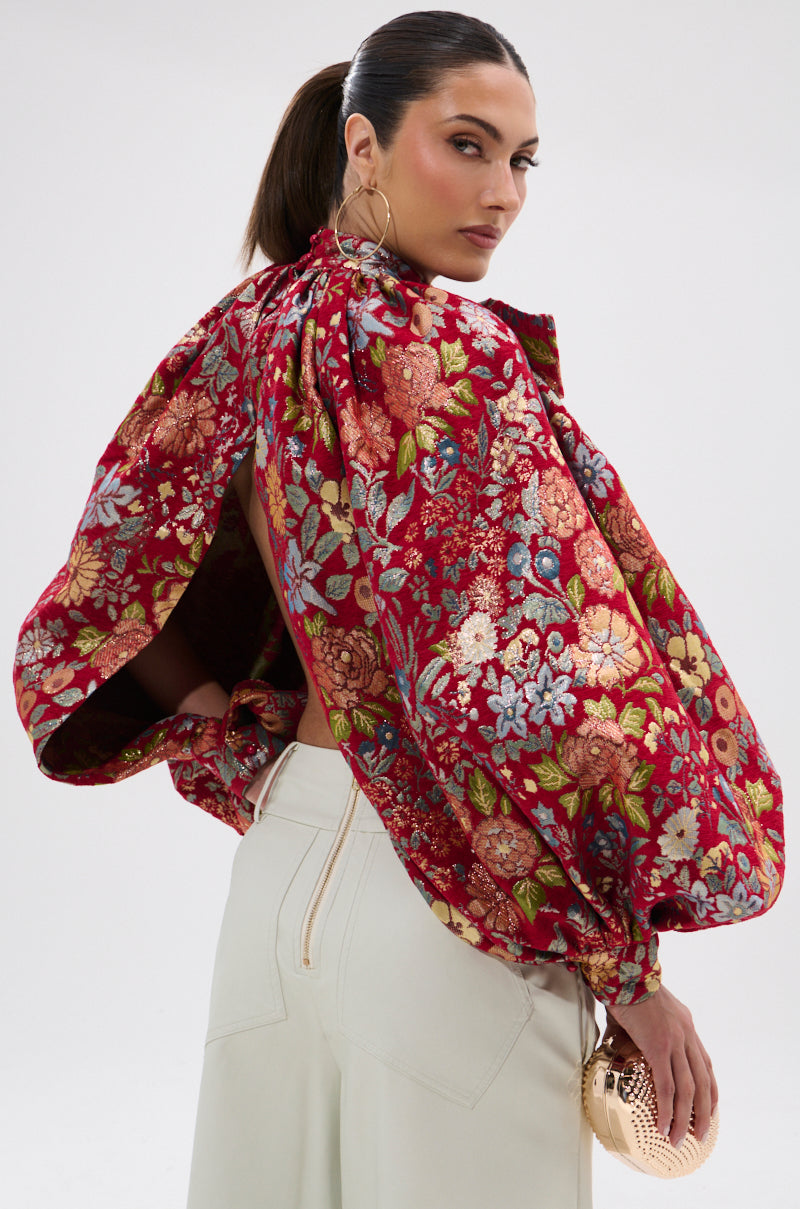 A woman with slicked-back hair wears the ETHEREAL FLORAL PUFF SLEEVE BROCADE TOP and high-waisted white pants, holding a small gold clutch. She looks over her shoulder against a plain background.