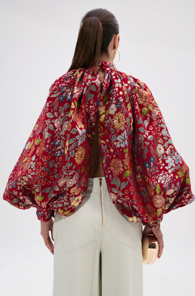 A woman with long brown hair in a ponytail wears the ETHEREAL FLORAL PUFF SLEEVE BROCADE TOP, featuring balloon sleeves and an open back, paired with cream high-waisted pants, a metallic clutch, and large gold hoop earrings.