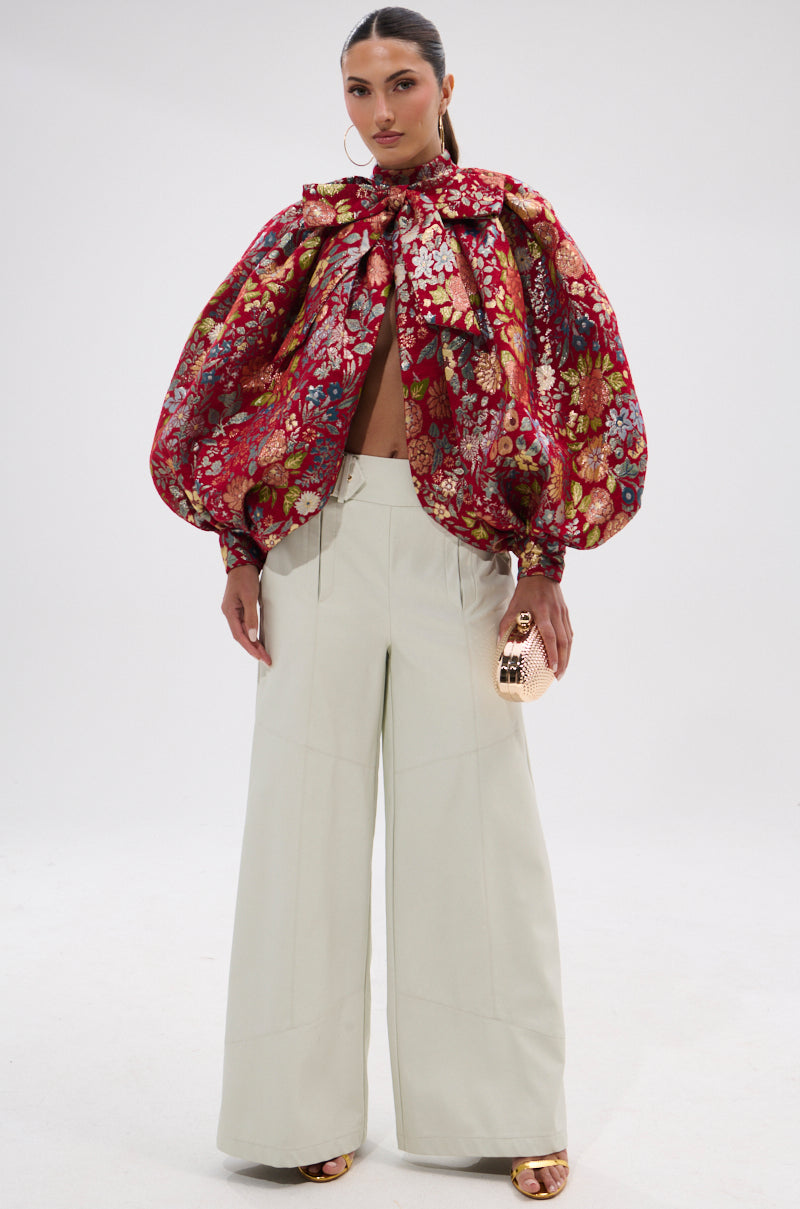 A woman models the ETHEREAL FLORAL PUFF SLEEVE BROCADE TOP with a bow at the neck, styled with wide-leg white pants, a metallic gold mini bag, and gold sandals against a plain background.