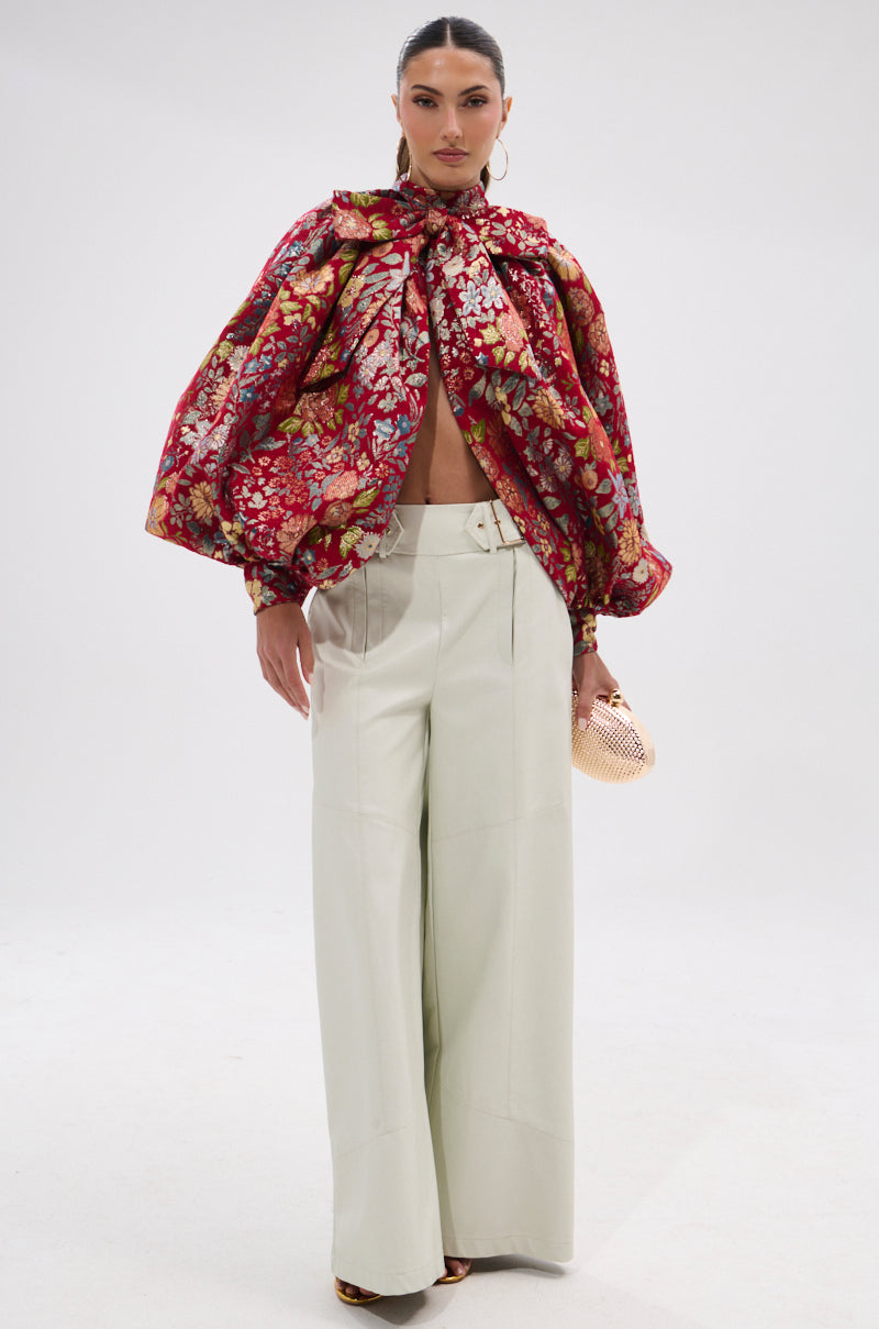 A woman in the ETHEREAL FLORAL PUFF SLEEVE BROCADE TOP and high-waisted wide-leg white trousers stands against a plain white backdrop, holding a small gold clutch with subtle metallic detailing.