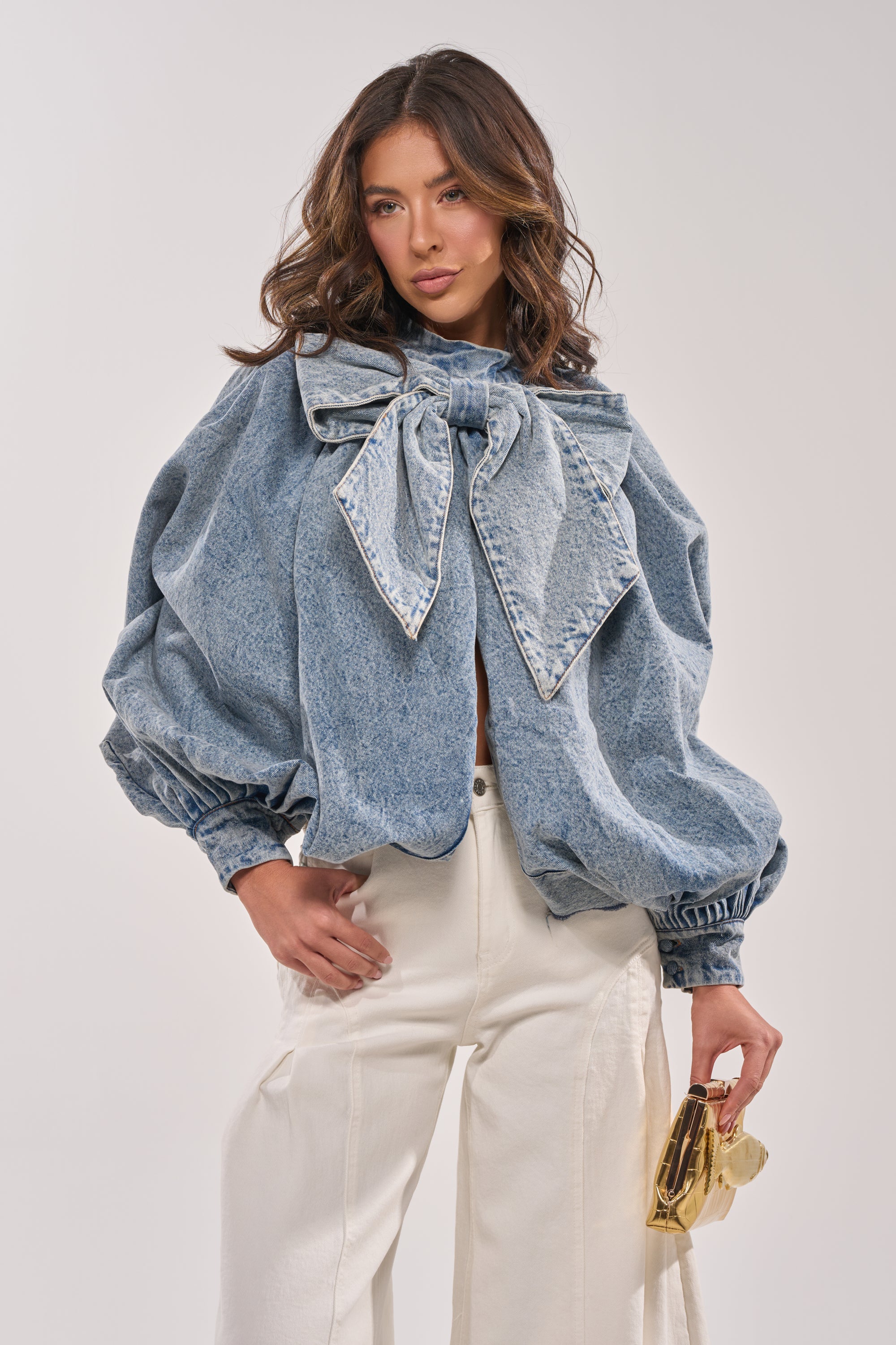 A woman models the NEW DREAM PUFF SLEEVE TOP, a light-wash denim piece with an oversized collar bow and billowy sleeves, paired with high-waisted white pants. She carries a small gold handbag and has loose, wavy hair. 2