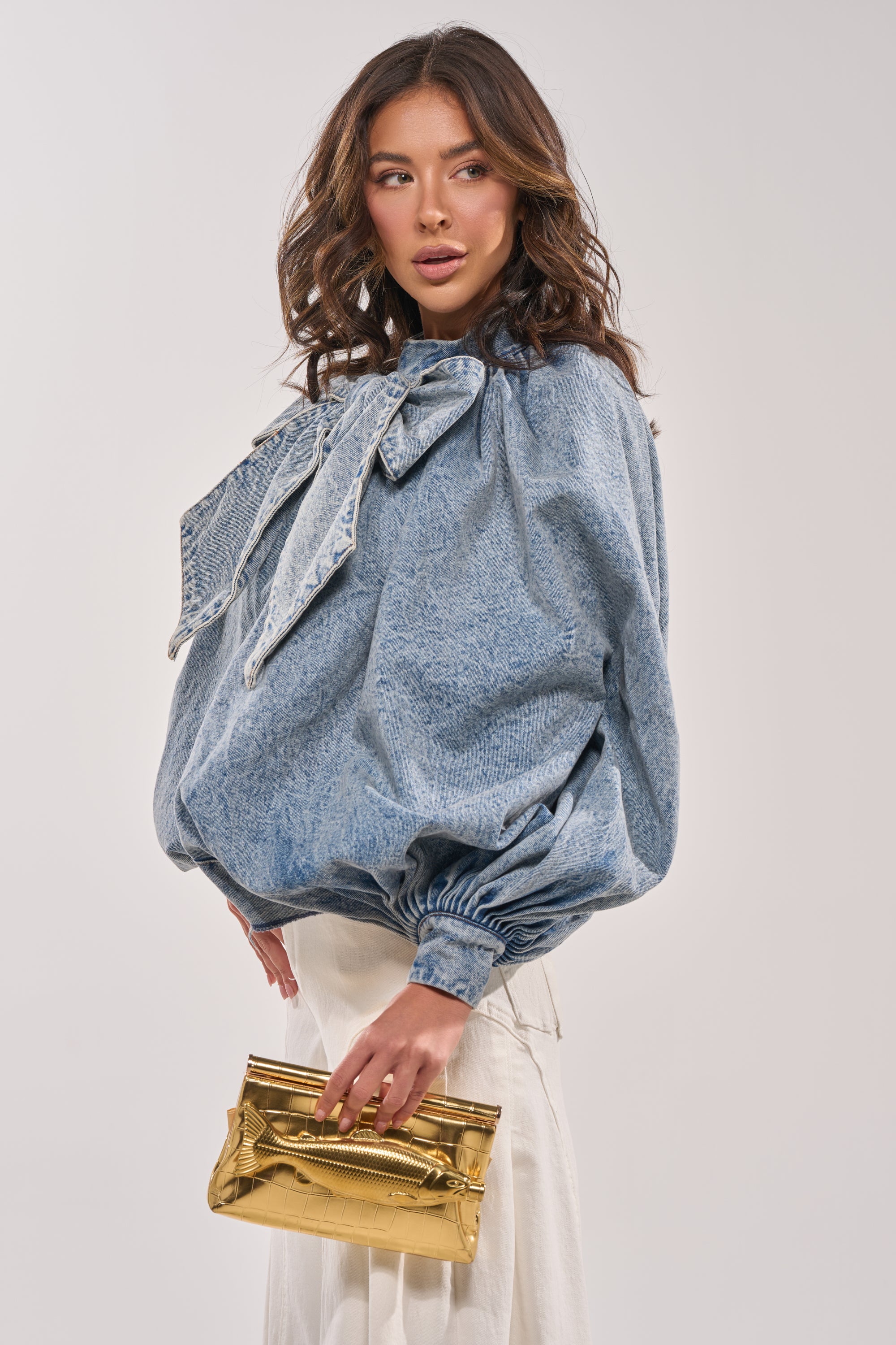 A woman models the NEW DREAM PUFF SLEEVE TOP—a statement denim bow design with puffy sleeves—paired with white pants and a shiny gold clutch, set against a plain light background. 5