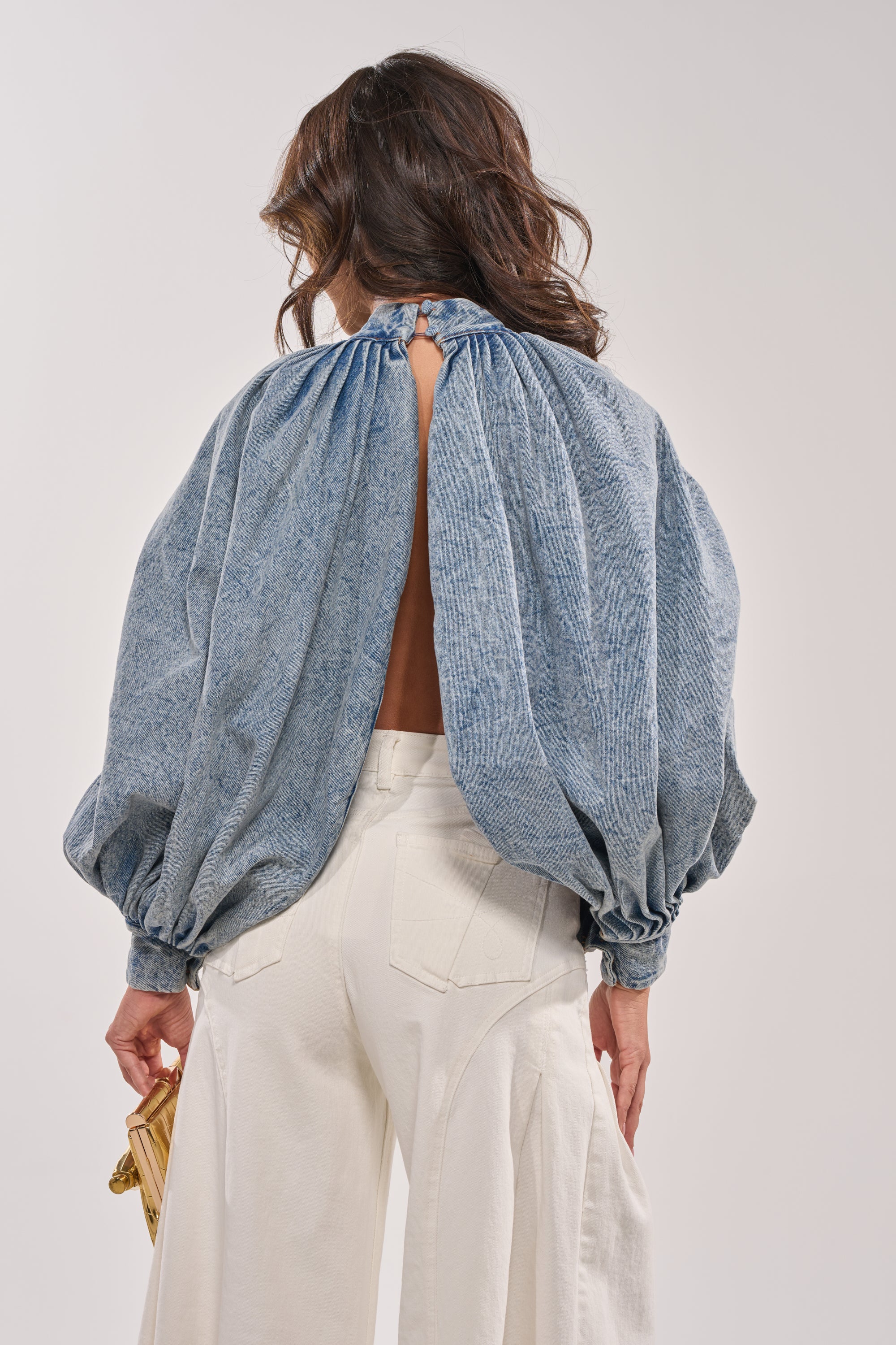 A woman is seen from the back wearing the NEW DREAM PUFF SLEEVE TOP in denim with voluminous sleeves, paired with white wide-legged pants and holding a small beige woven clutch in her left hand. 6