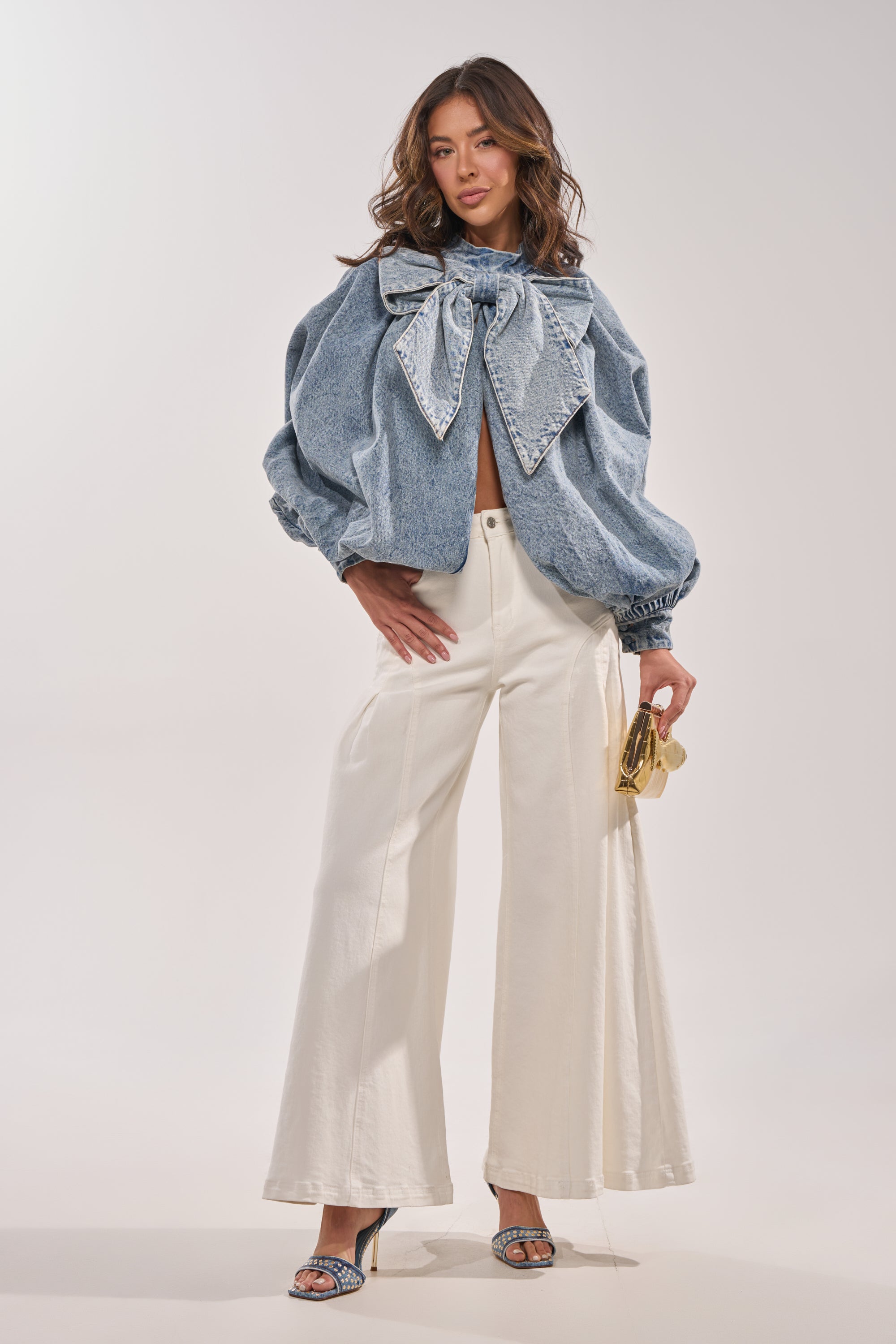A woman models the NEW DREAM PUFF SLEEVE TOP with an oversized bow, paired with wide-leg white pants, high-heeled sandals, and a small gold handbag against a plain light background. 7