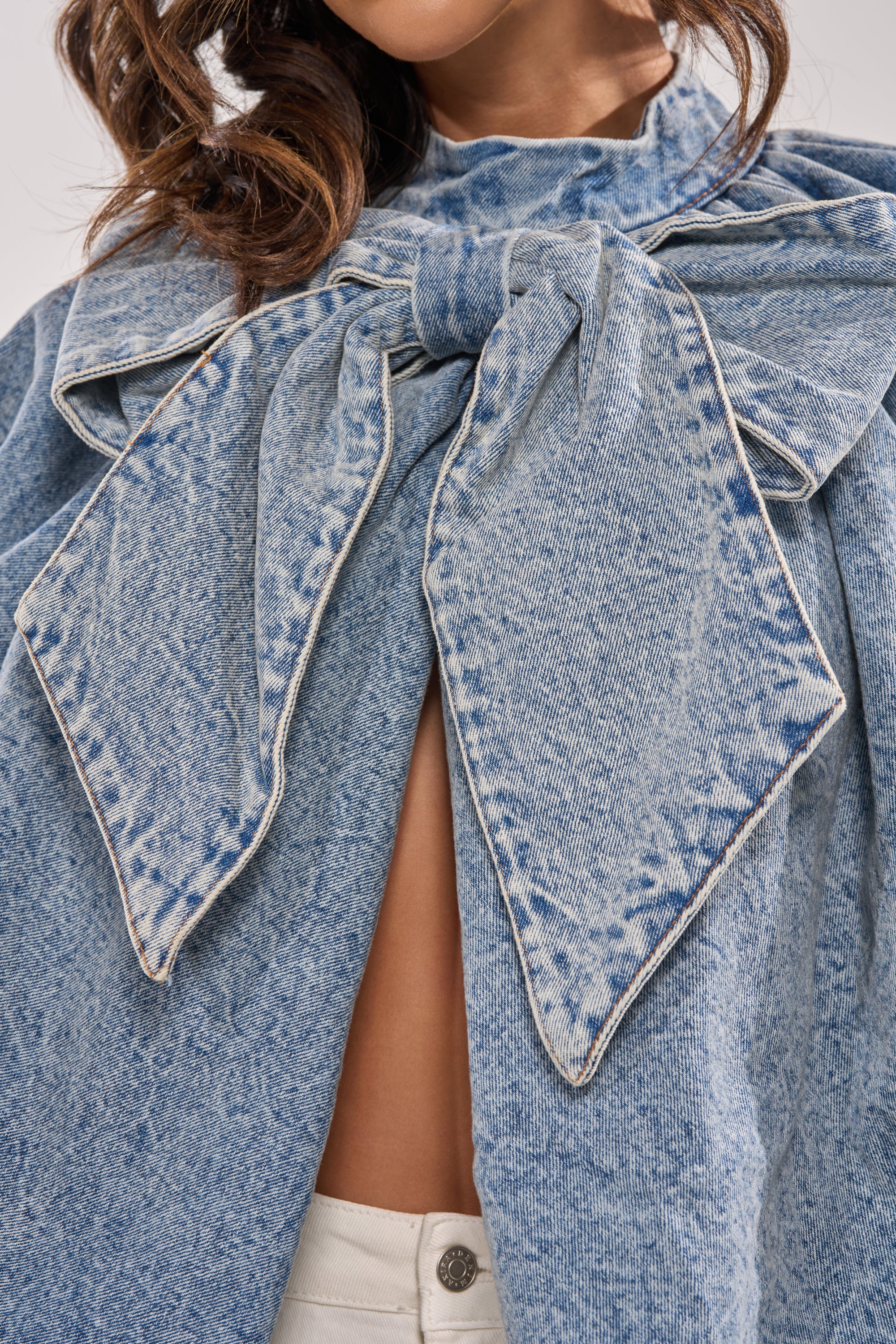 A person wears the NEW DREAM PUFF SLEEVE TOP, a statement denim piece with a large, faded bow covering the chest and upper torso, paired with white pants. The look is bold and modern, with the face partially out of frame. 8