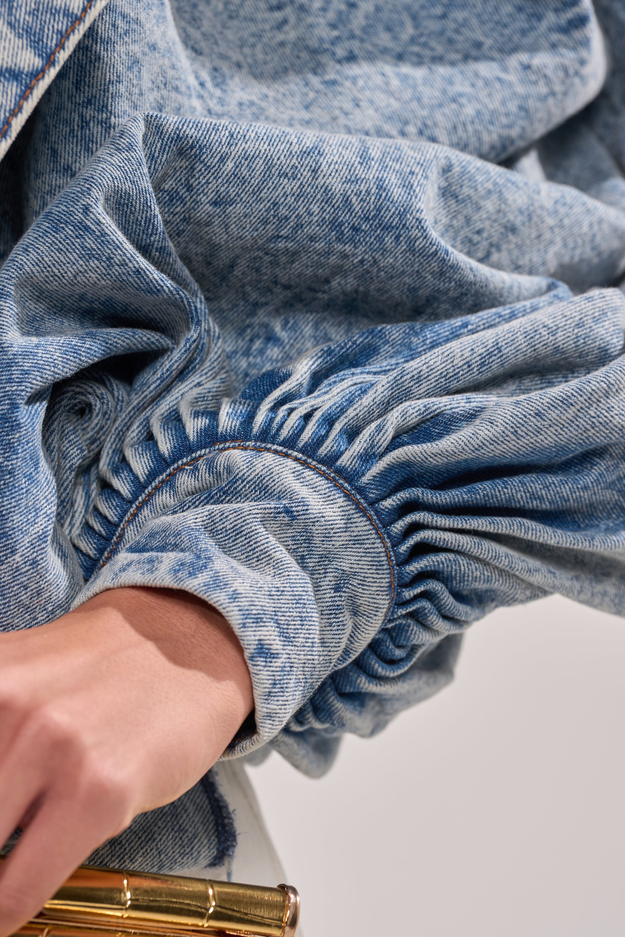 Close-up of a hand holding a gold object, with the arm wearing the NEW DREAM PUFF SLEEVE TOP—a light blue denim top featuring distinctive gathered sleeves. 9