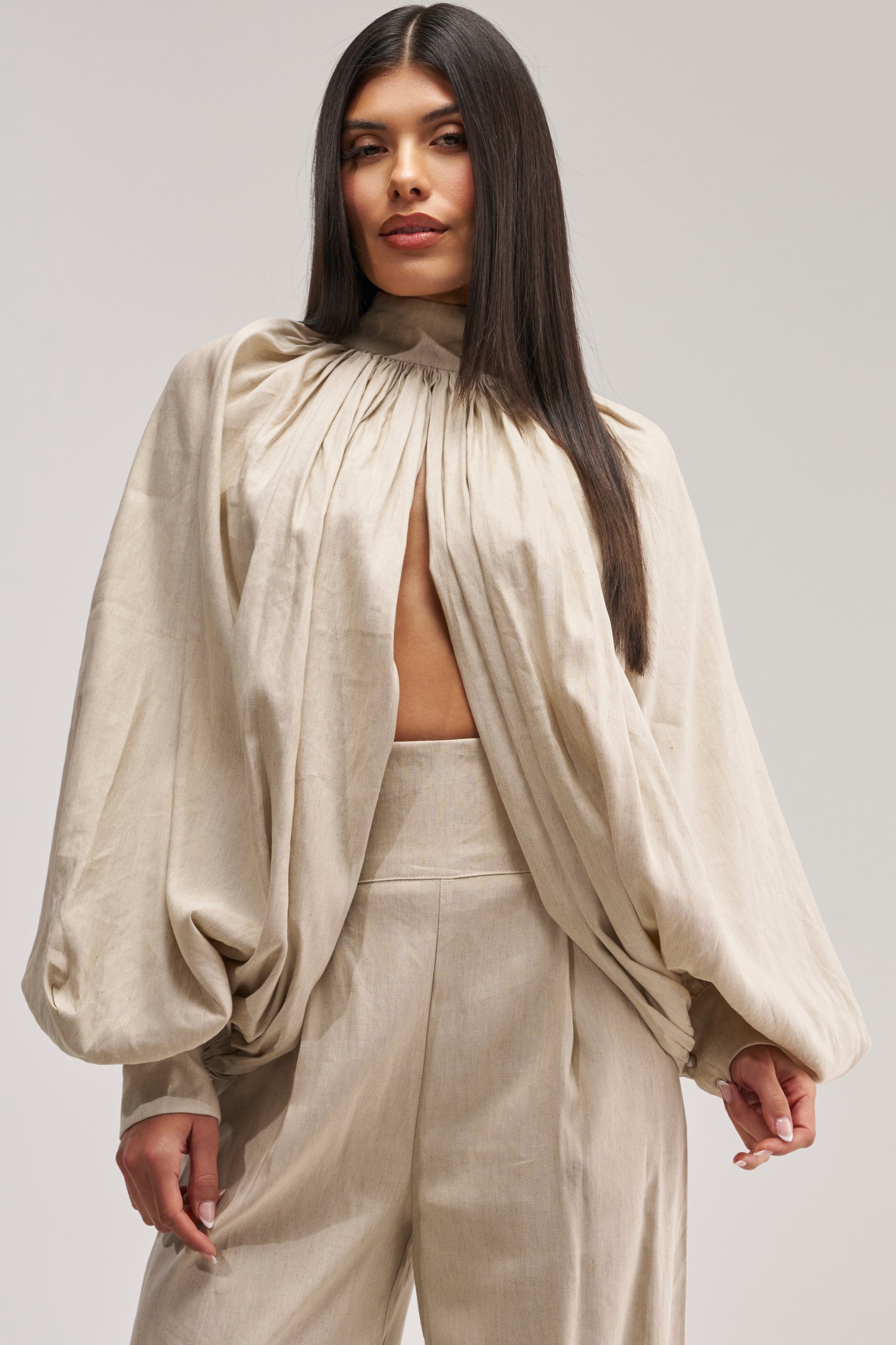 Wearing the MASTERPIECE PUFF LONG SLEEVE TOP IN LINEN—a loose, beige linen blouse with a ruched neckline—paired with wide-leg trousers, this woman exudes effortless chic. Her straight, dark hair is parted in the middle as she stands against a plain light background, creating a look that's perfect for a summer date night.