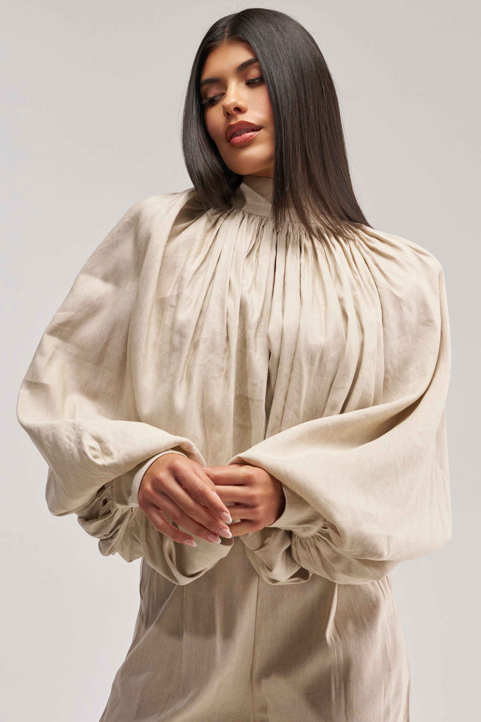 A woman with long, straight dark hair wears the MASTERPIECE PUFF LONG SLEEVE TOP IN LINEN—a loose, light beige blouse featuring pleated details and voluminous puff sleeves—standing against a plain background, creating the perfect look for a summer date night.