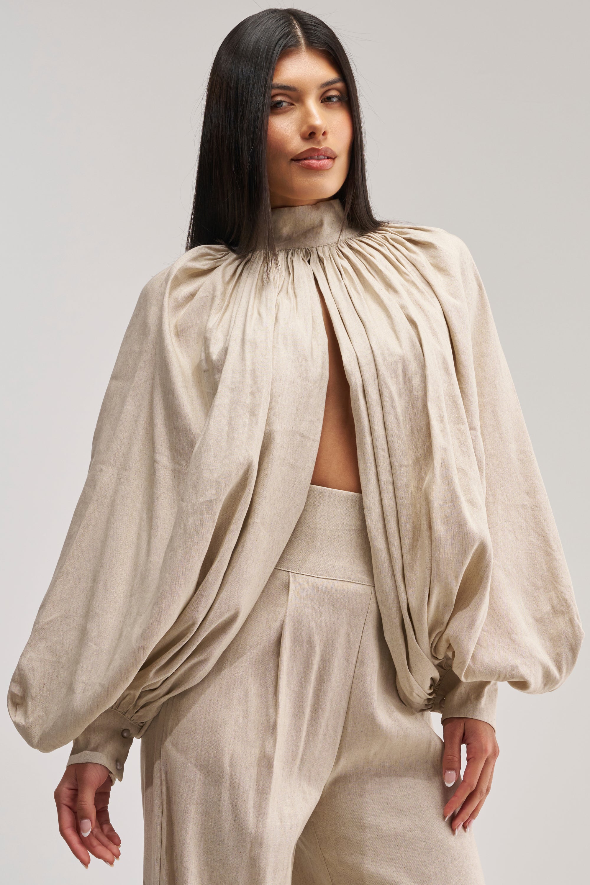 Clad in the MASTERPIECE PUFF LONG SLEEVE TOP IN LINEN—a beige high-neck blouse featuring dramatic billowy puff sleeves and a front slit—she pairs it with matching high-waisted beige pants. With her long, straight dark hair and minimalist background, she exudes an effortlessly chic vibe, making this look ideal for a summer date night.