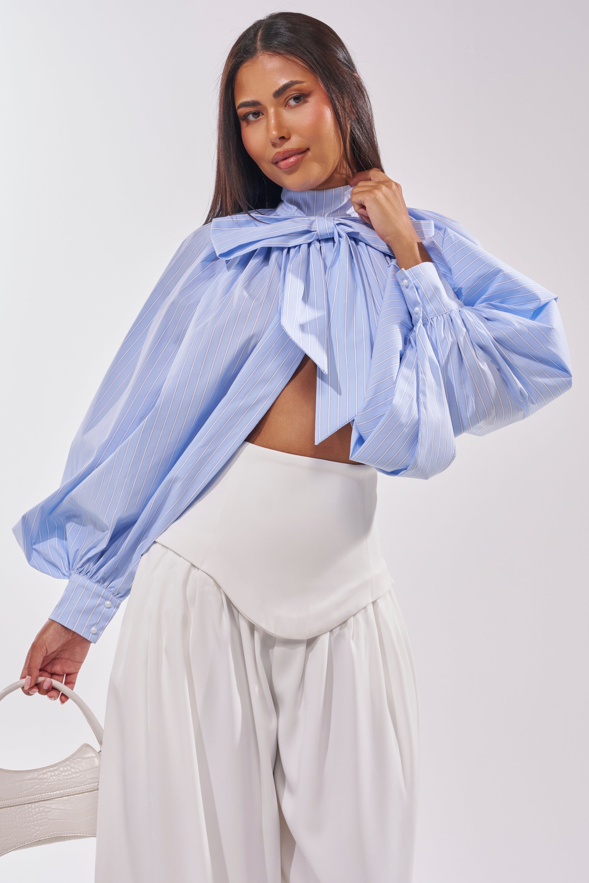 A woman wears the MASTERPIECE POPLIN PUFF LONG SLEEVE TOP IN BLUE with high-waisted white trousers and holds a small white handbag. 0