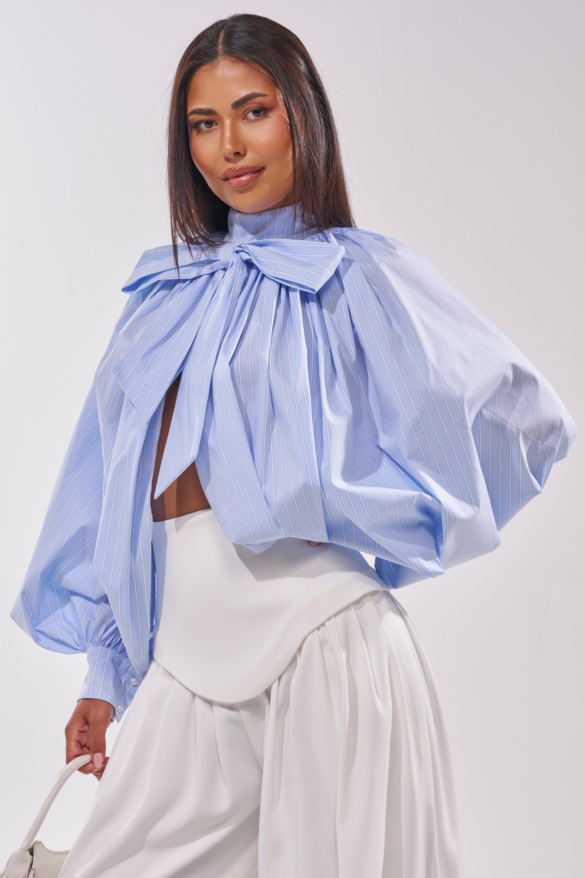 A woman stands against a plain light background, wearing the MASTERPIECE POPLIN PUFF LONG SLEEVE TOP IN BLUE with an oversized bow neckline and billowy sleeves, paired with white high-waisted, wide-leg pants. 1