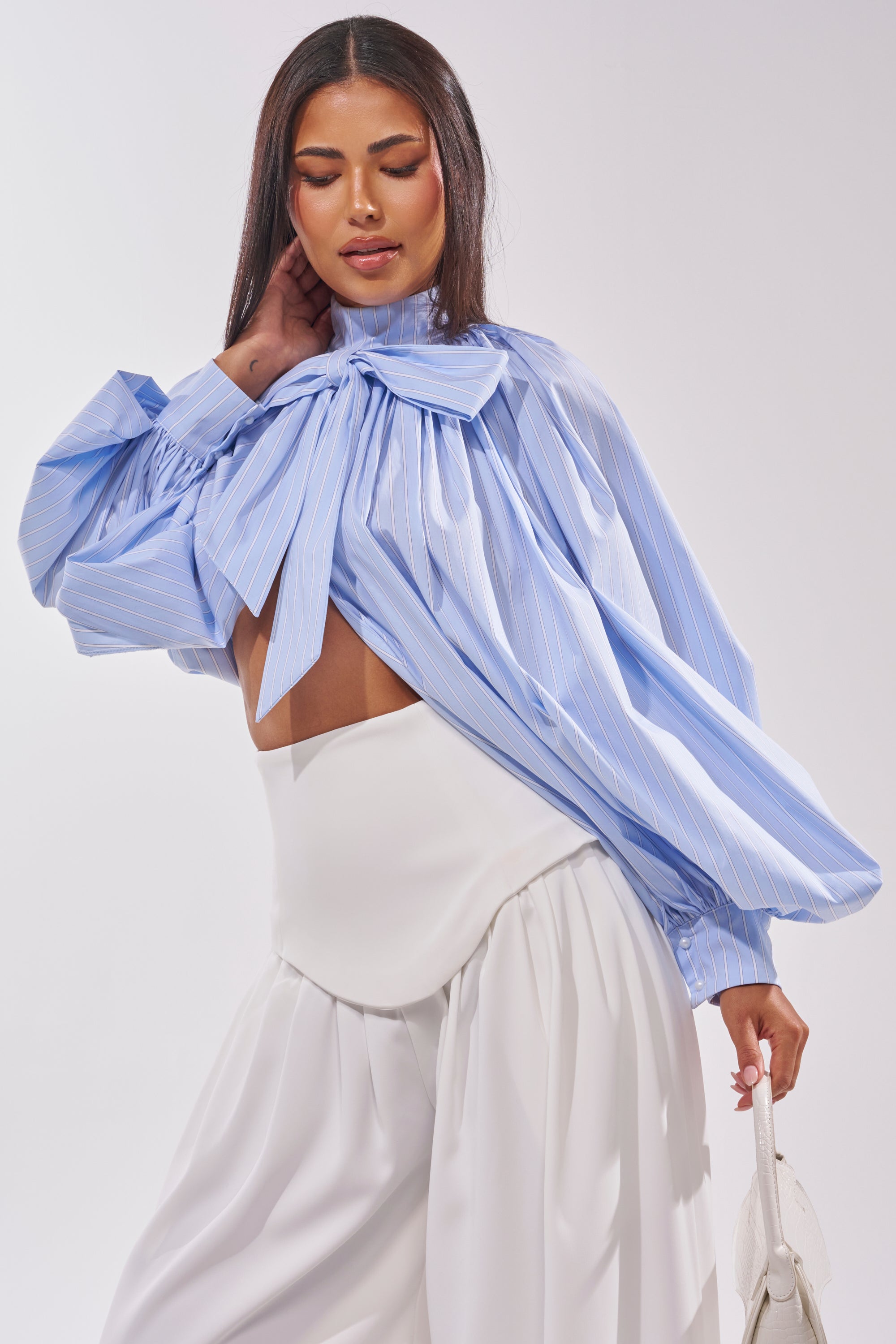 A woman models the MASTERPIECE Poplin Puff Long Sleeve Top in blue, featuring stripes and an oversized bow neckline, styled with high-waisted white flowy pants and a white handbag. 2