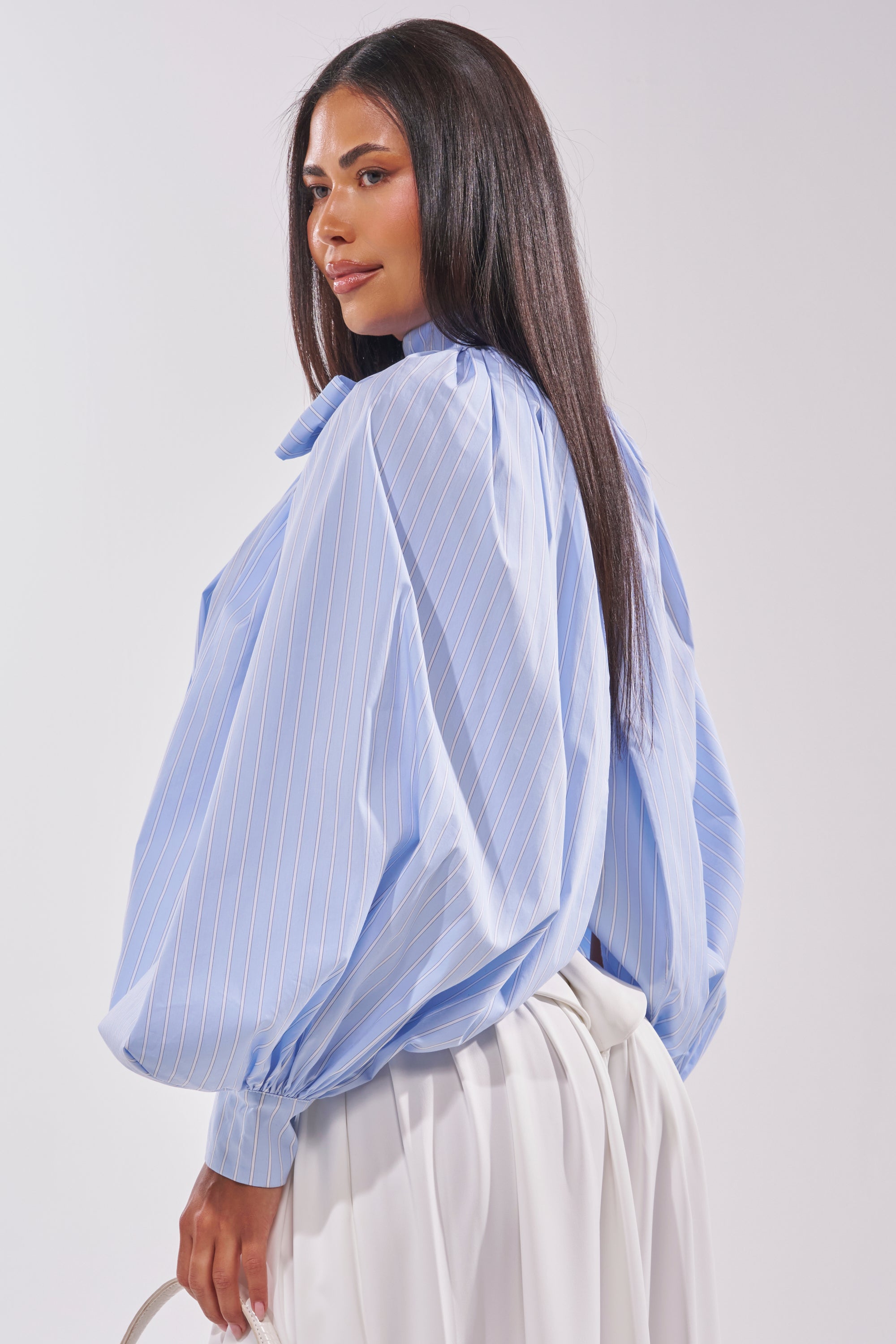 A woman with straight, dark hair wears the MASTERPIECE POPLIN PUFF LONG SLEEVE TOP IN BLUE and white pleated pants, posing against a light background. 3
