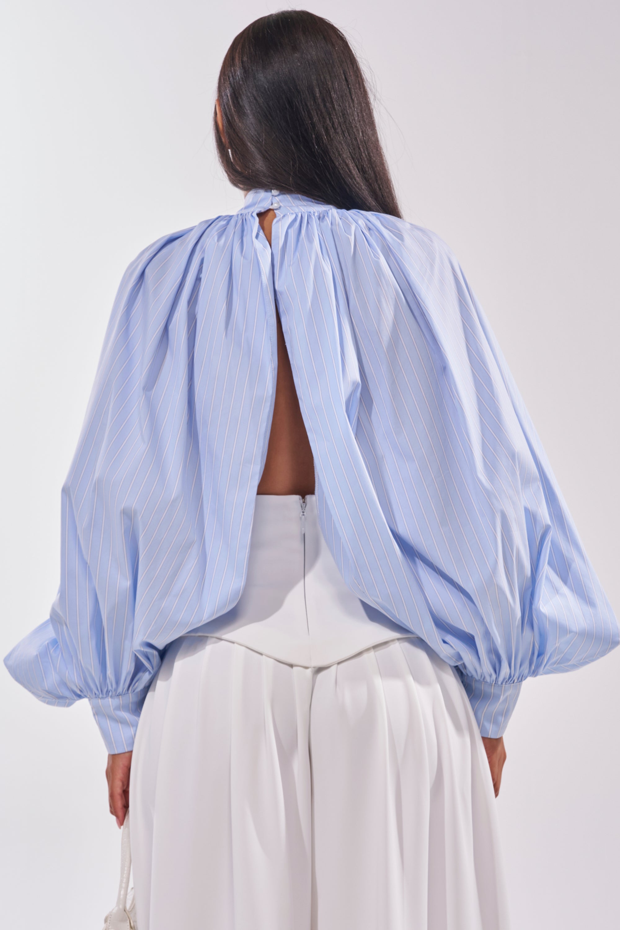 A person with long dark hair is seen from behind wearing the MASTERPIECE POPLIN PUFF LONG SLEEVE TOP IN BLUE, featuring light blue pinstripes, a keyhole open back, and balloon sleeves, paired with a high-waisted white skirt. 4
