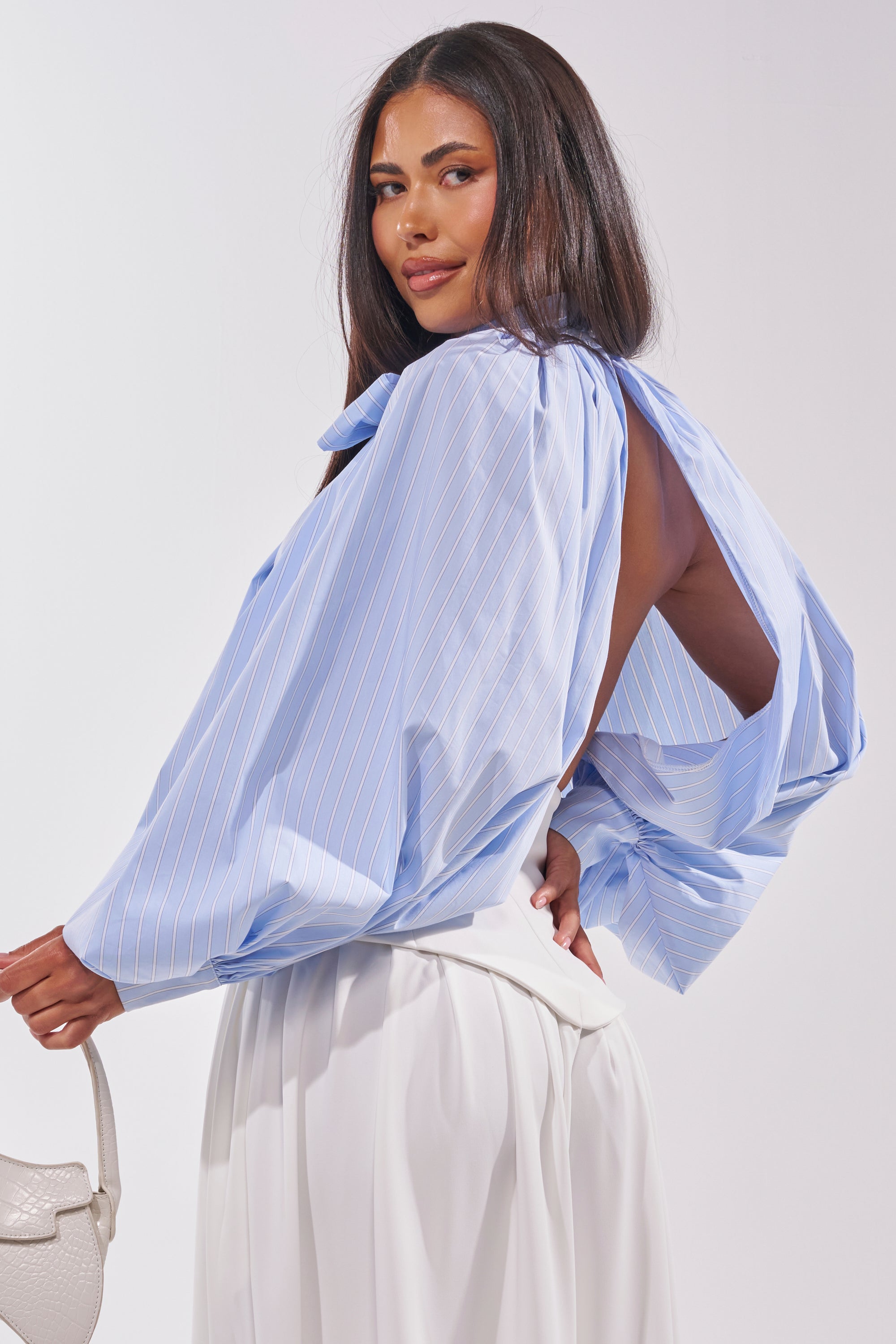 A woman with long dark hair smiles over her shoulder, wearing the MASTERPIECE POPLIN PUFF LONG SLEEVE TOP IN BLUE—featuring balloon sleeves and white pinstripes—paired with white high-waisted pants and a white handbag. 5