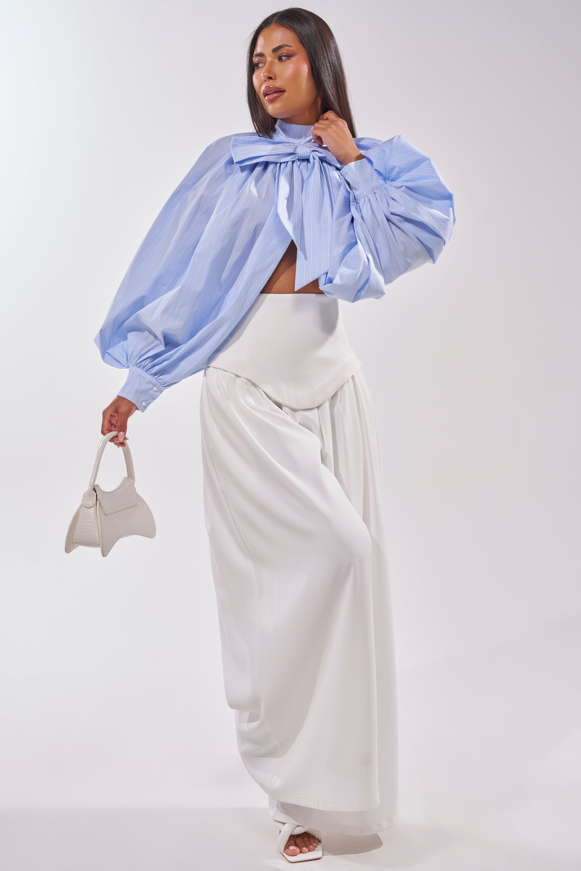 A woman confidently poses in the MASTERPIECE POPLIN PUFF LONG SLEEVE TOP IN BLUE with blue stripes, balloon sleeves, and an oversized bow neckline, styled with high-waisted wide-leg white pants, a white handbag, and heels. 7