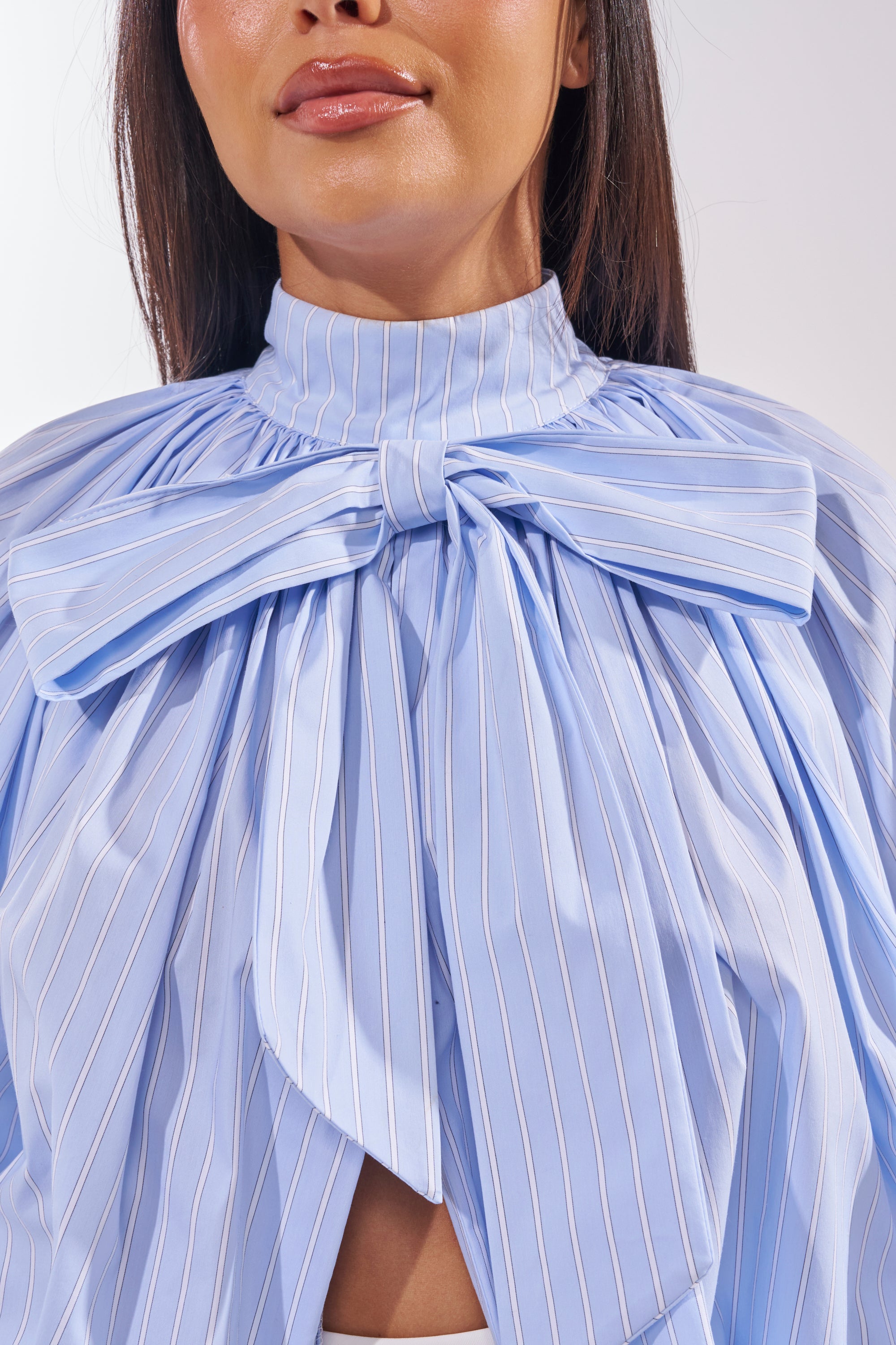 A woman wears the MASTERPIECE POPLIN PUFF LONG SLEEVE TOP IN BLUE, a pinstriped blouse with a high neck and oversized bow. Her long dark hair is straight, showing only the lower half of her face. 8
