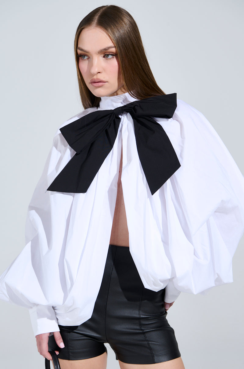 A person with long brown hair wears the BLACK TIE AFFAIR BLOUSE, featuring peasant-style sleeves and a large black bow at the mock neck. Paired with black leather shorts and a small black bag, they stand against a plain gray backdrop.