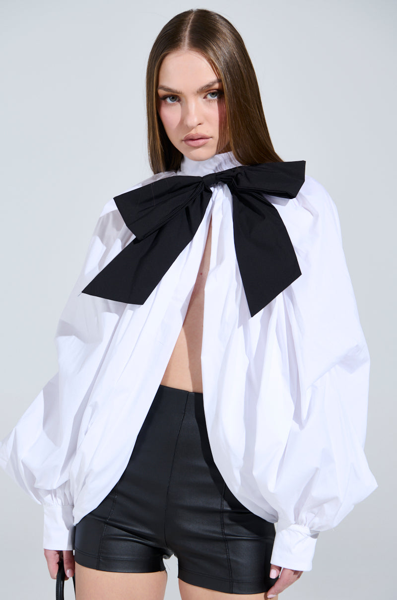 A person with long hair wears the BLACK TIE AFFAIR BLOUSE with peasant-style sleeves and a large black neck bow, paired with black leather shorts on a plain light gray background.