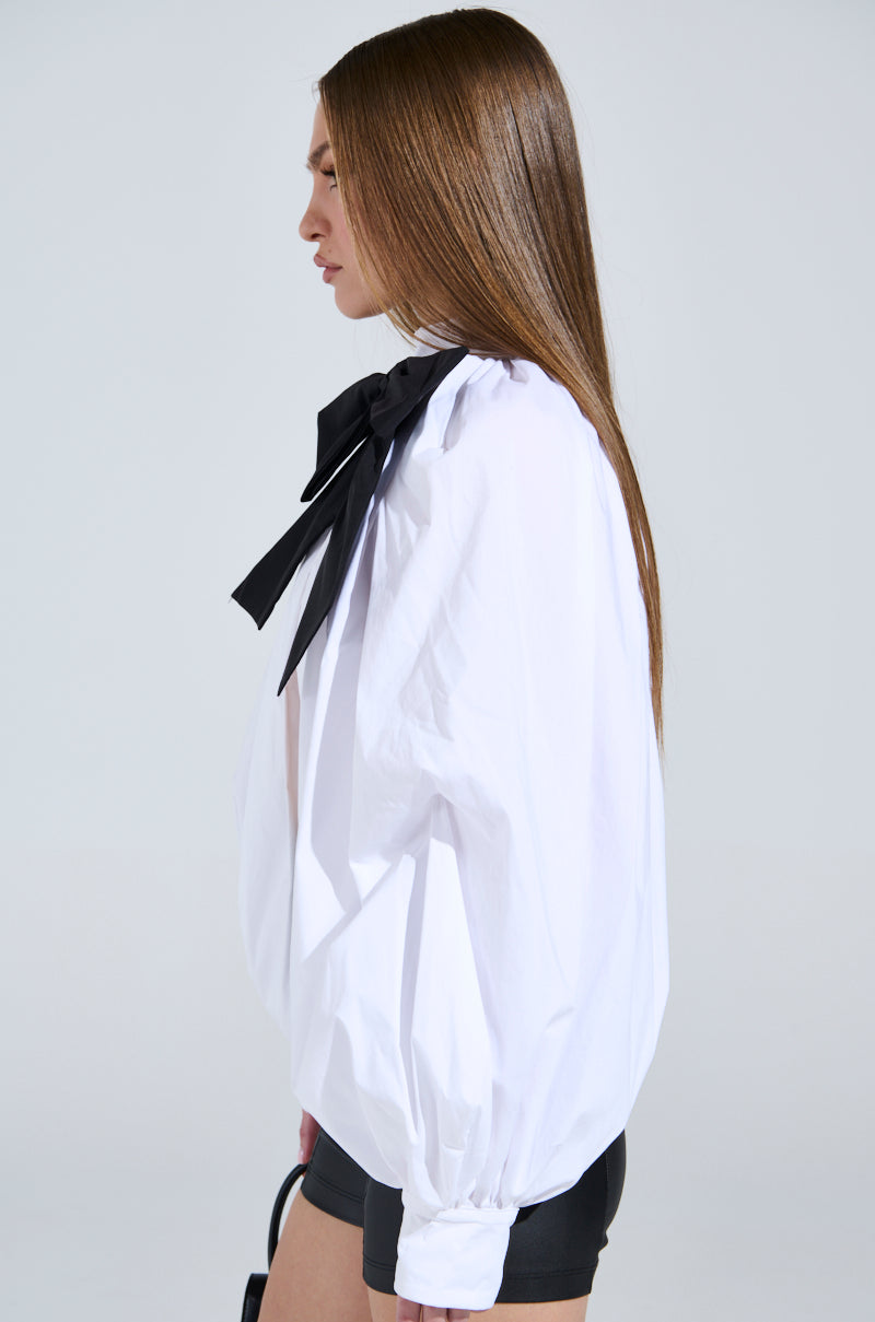 With long, straight hair, the individual stuns in the white BLACK TIE AFFAIR BLOUSE with elegant peasant sleeves and a bold black bow. They pair it with black shorts and hold a small handbag, facing left against a plain backdrop.