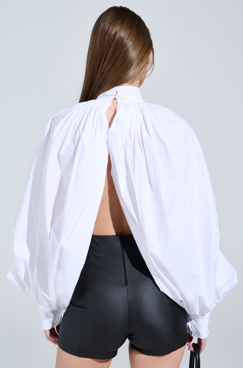 A person with long hair faces away, highlighting the BLACK TIE AFFAIR BLOUSE with peasant-style sleeves and an open back, matched with black high-waisted shorts, set against a plain gray backdrop.