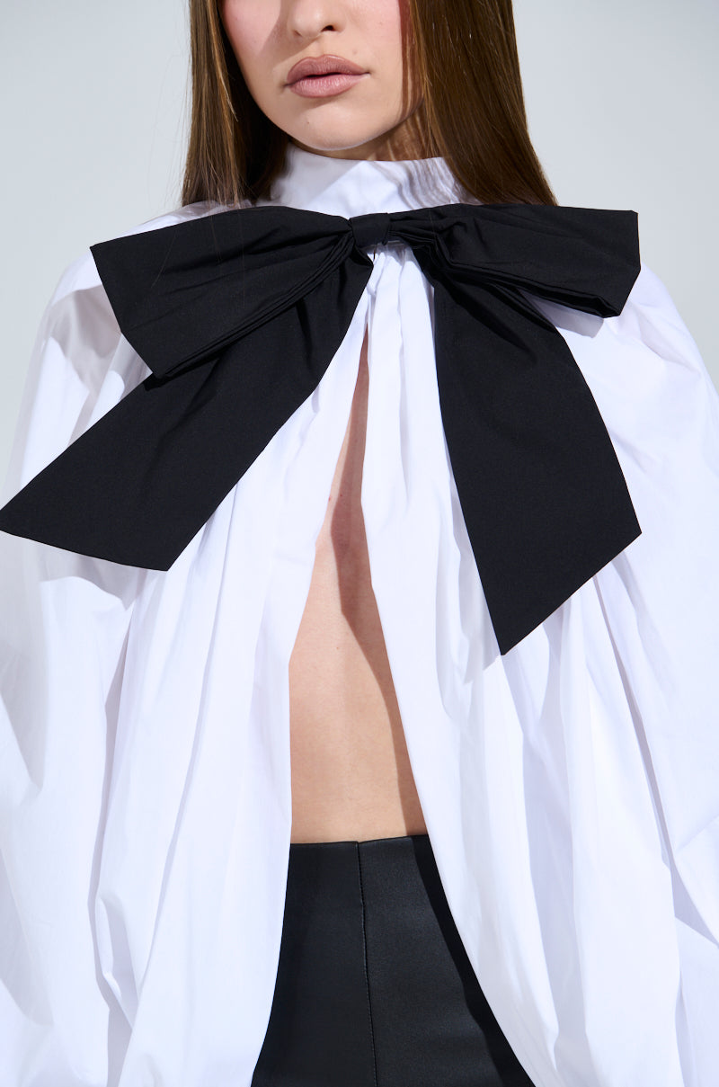 A person sports the BLACK TIE AFFAIR BLOUSE, a white peasant-style top with an oversized black neck bow and wide open front, paired with black pants. They have straight, long hair, and only part of their face is visible.