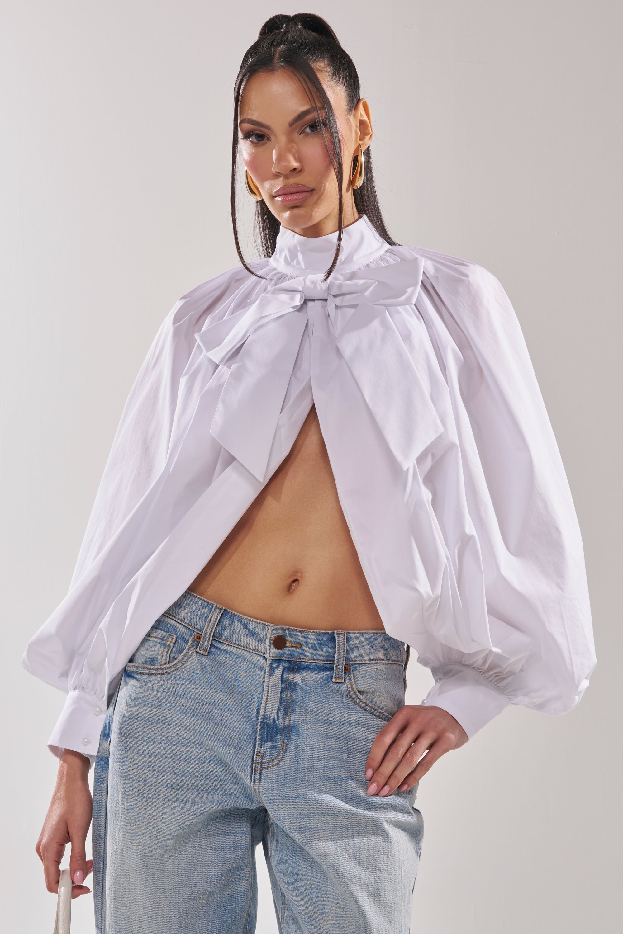A woman wears the BLACK TIE AFFAIR BLOUSE IN WHITE—a voluminous, open-front long sleeve blouse with puffed sleeves and a dramatic neck bow—styled with light blue jeans, hoop earrings, and her long straight hair loose. 0