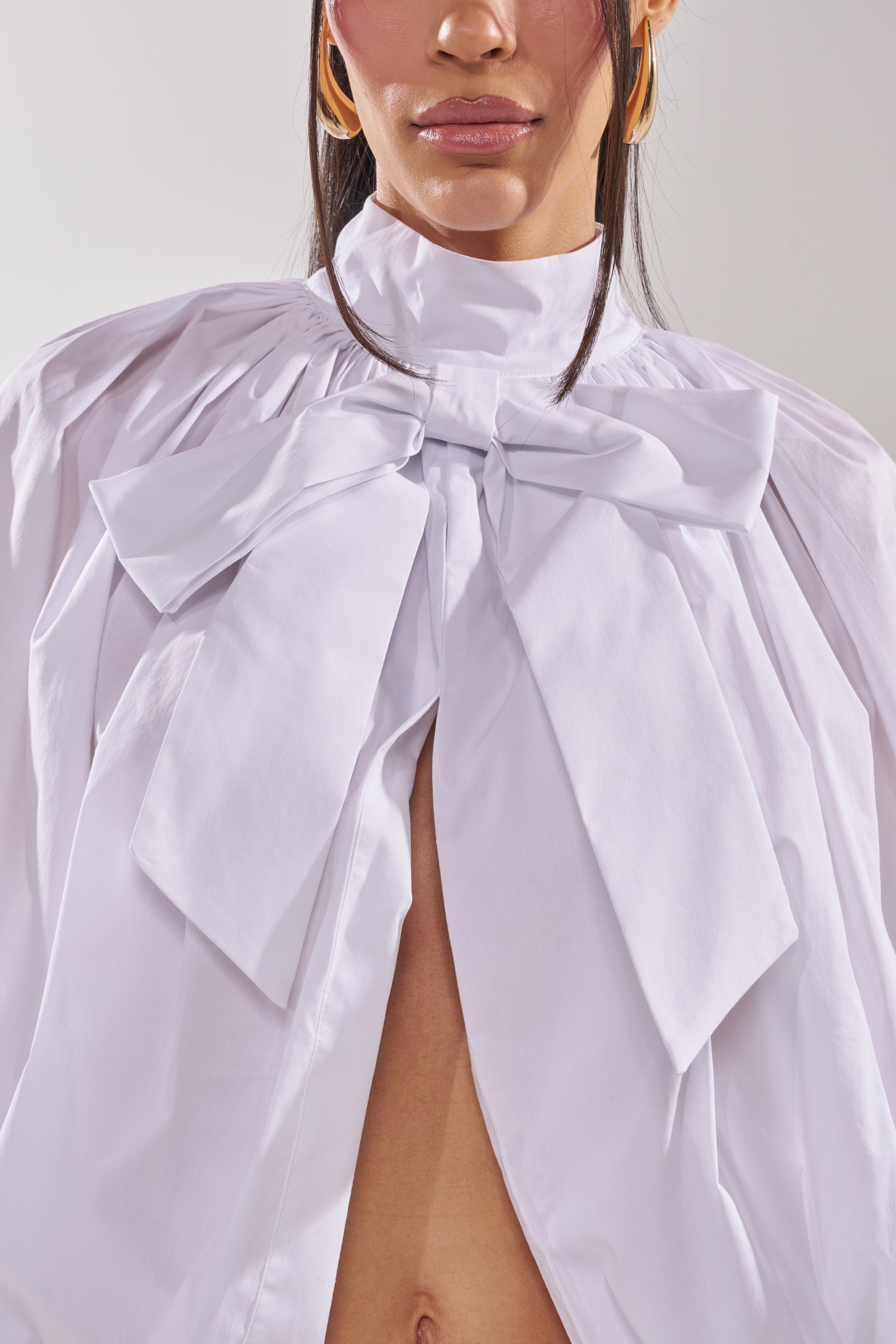 Wearing the BLACK TIE AFFAIR BLOUSE IN WHITE, a woman sports a high-collared statement blouse with an oversized bow, open below the neck to reveal her midriff. She pairs voluminous sleeves with gold hoops and subtle makeup. 9