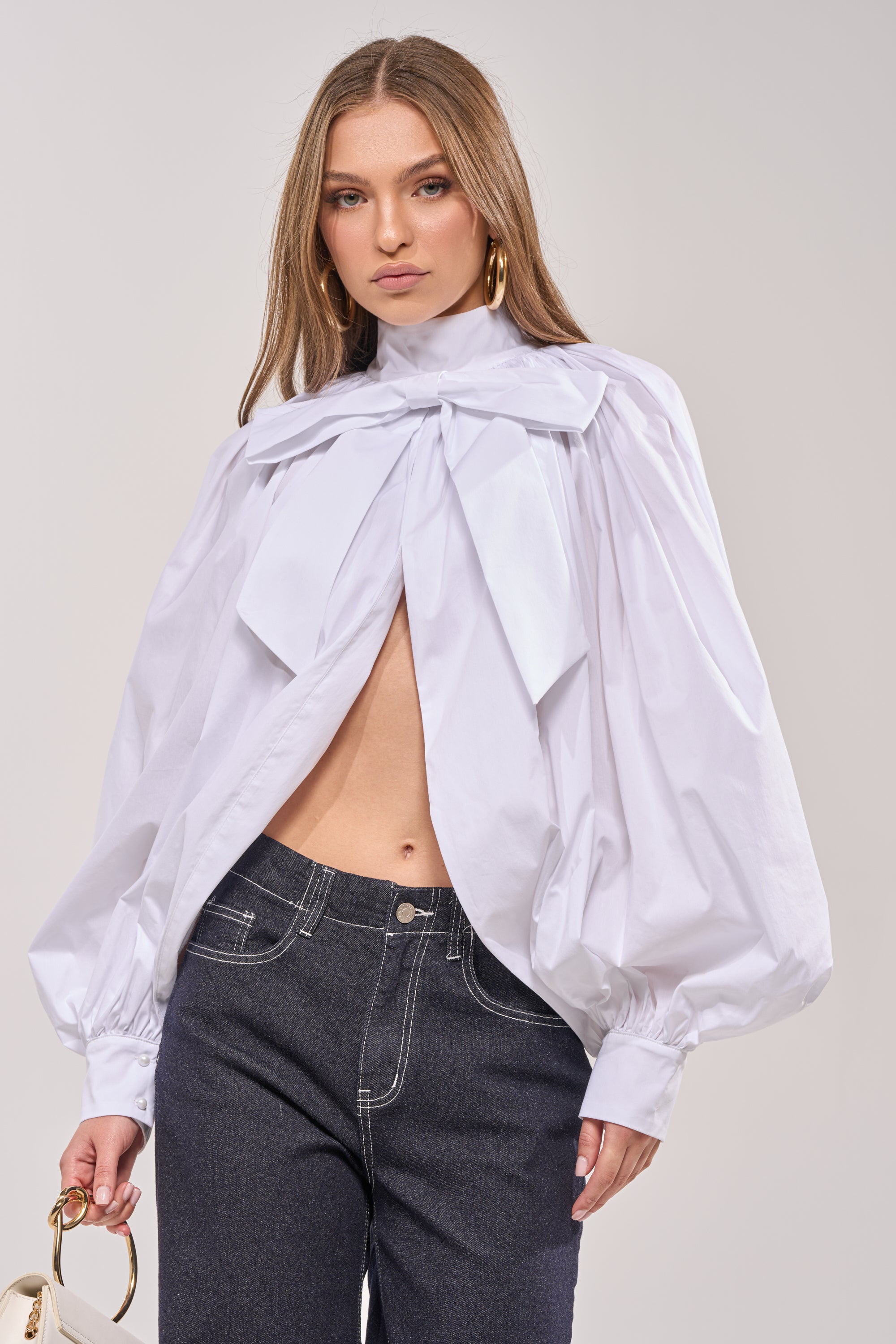Wearing the BLACK TIE AFFAIR BLOUSE IN WHITE with voluminous sleeves, an oversized bow-tie neckline, and front cut-out, a woman pairs it with high-waisted dark jeans and holds a small handbag against a plain light background. 0