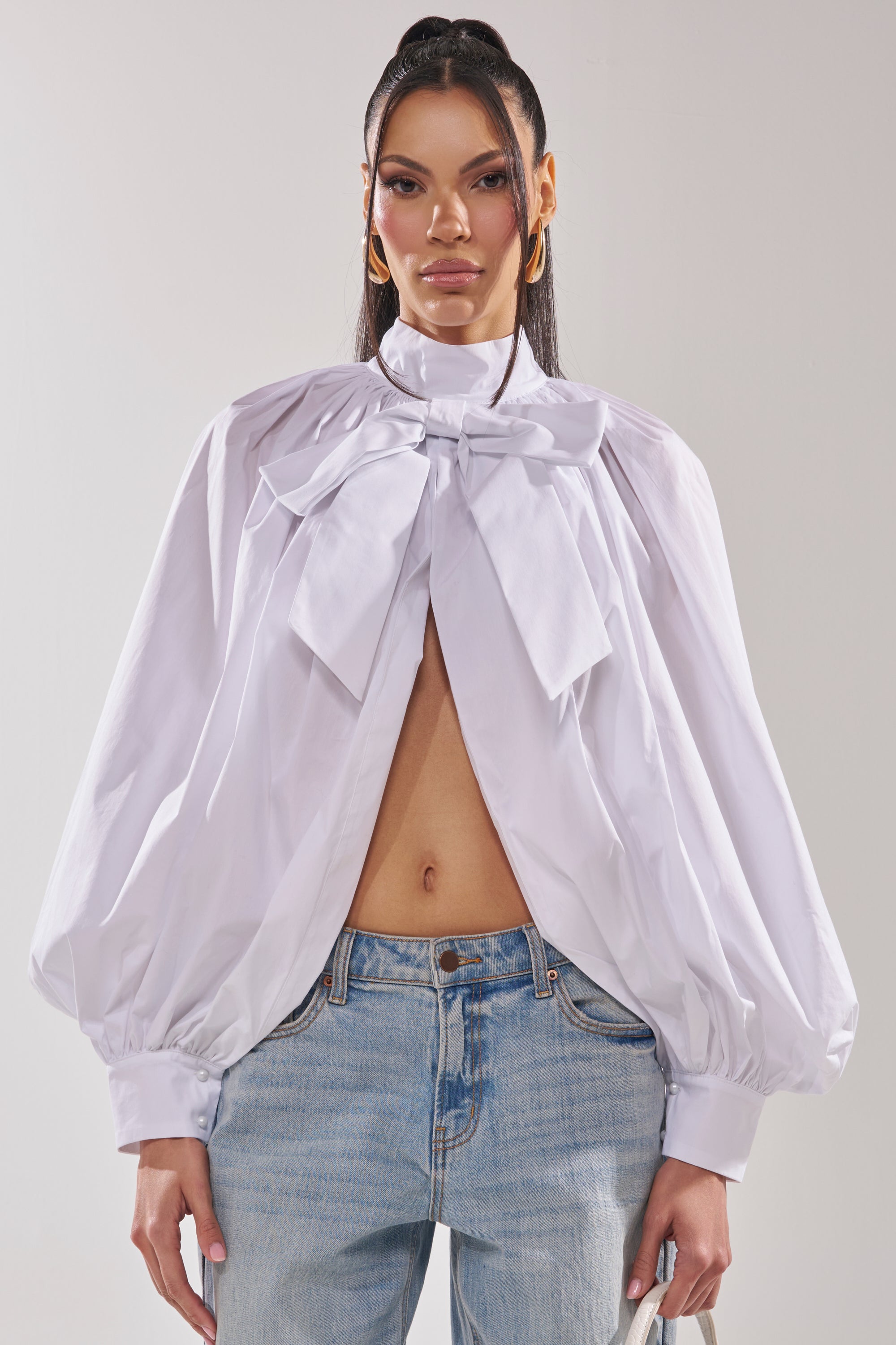 A woman wears the BLACK TIE AFFAIR BLOUSE IN WHITE, featuring a large neck bow and billowy sleeves, paired with light blue jeans, standing against a plain background. 1