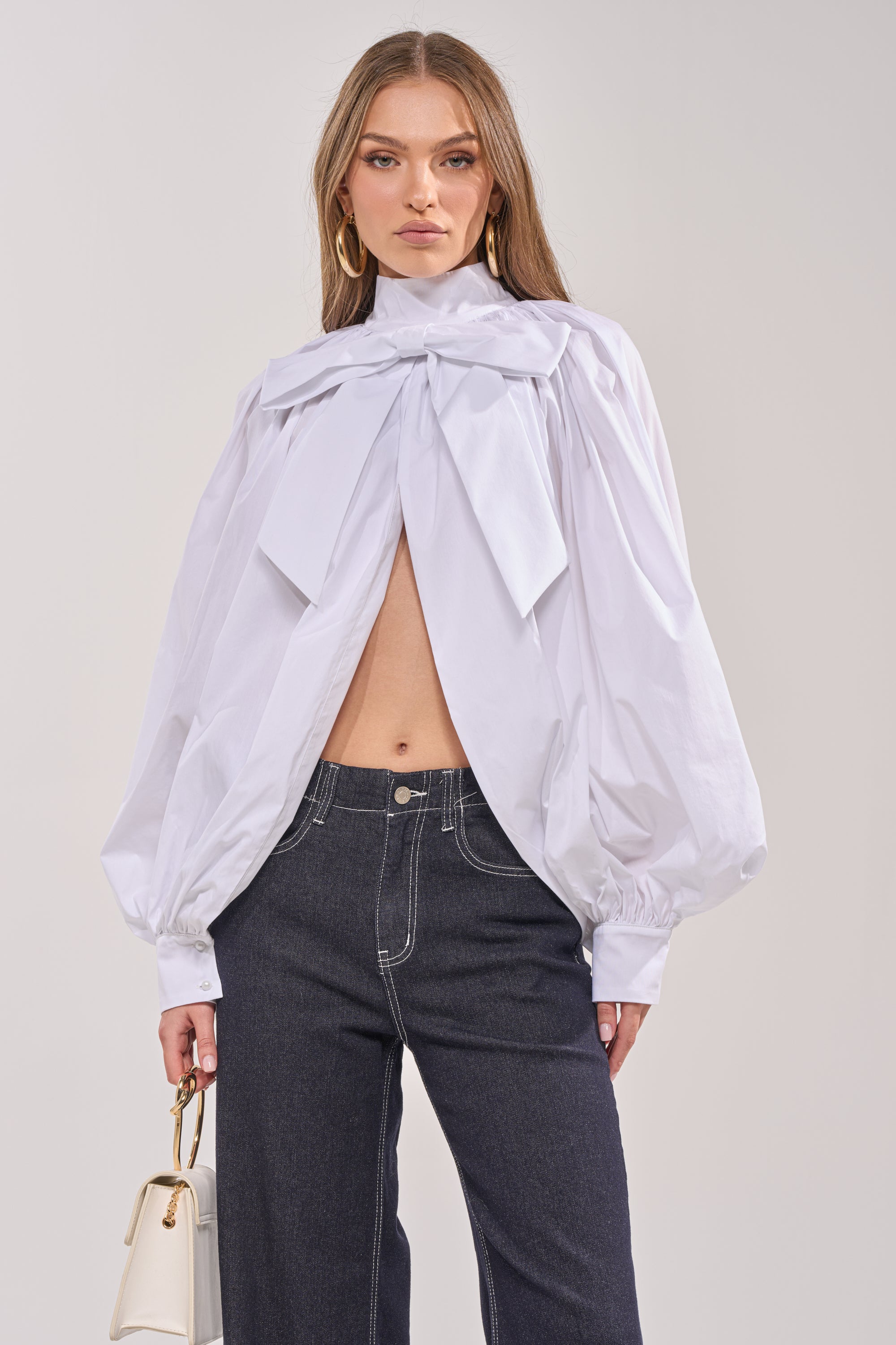 Wearing the BLACK TIE AFFAIR BLOUSE IN WHITE with an oversized bow-tie neckline and puffy sleeves, they pair it with high-waisted dark jeans and a small white handbag, their long straight hair falling against a neutral background. 1