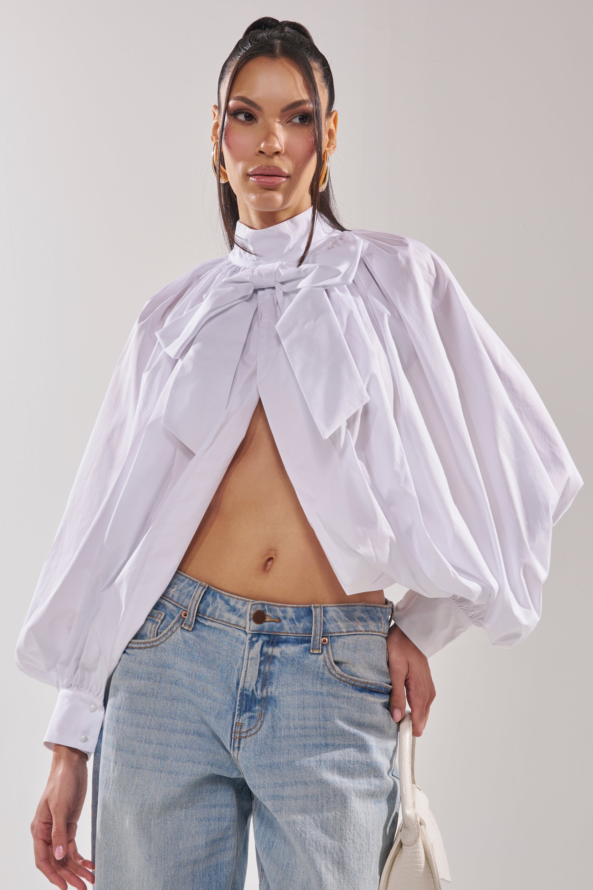 She wears the BLACK TIE AFFAIR BLOUSE IN WHITE with voluminous long sleeves and a large bow, paired with light blue jeans. She holds a small white handbag and has her hair styled in a high ponytail. 2